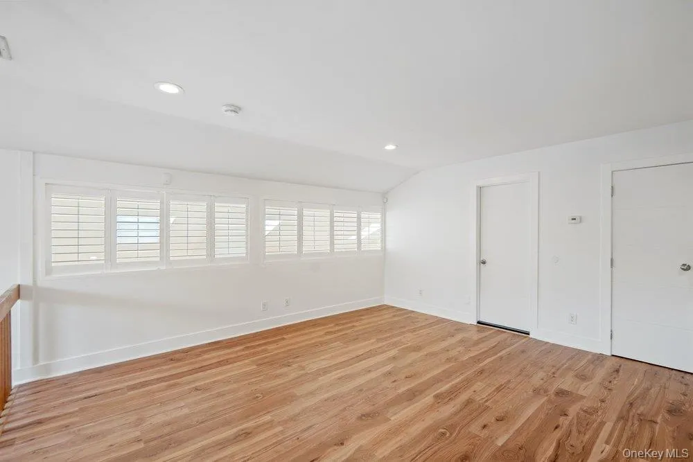 Vaulted ceiling, wood-finish flooring, recessed lighting, multiple windows with white plantation shutters, and two white panel doors Vaulted ceiling, wood-finish flooring, recessed lighting, multiple windows with white plantation shutters, and two white panel doors