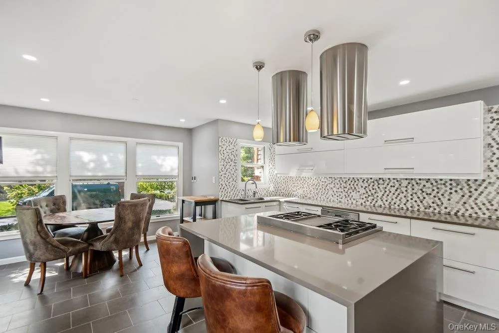 Kitchen featuring an island with an integrated cooktop, white flat-panel cabinetry, a mosaic tile backsplash, and sleek pendant lighting Kitchen featuring an island with an integrated cooktop, white flat-panel cabinetry, a mosaic tile backsplash, and sleek pendant lighting