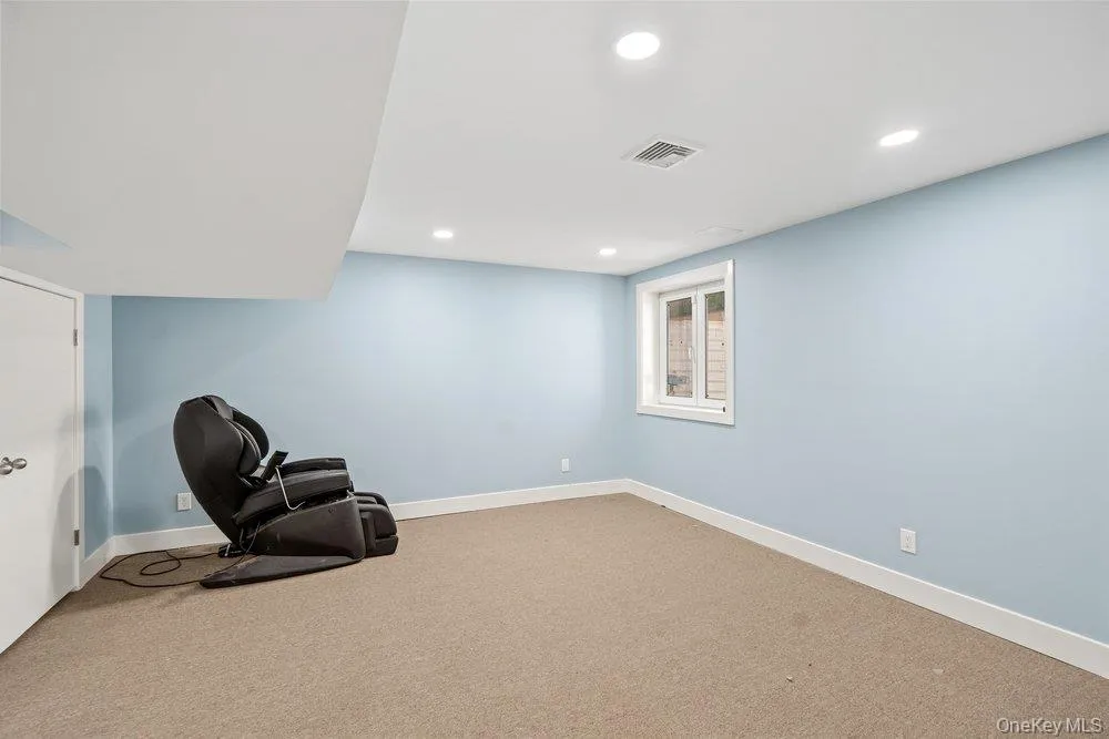 Spacious recreation room featuring soft blue painted walls, neutral carpet flooring, recessed ceiling lighting, and a large window allowing natural light Spacious recreation room featuring soft blue painted walls, neutral carpet flooring, recessed ceiling lighting, and a large window allowing natural light
