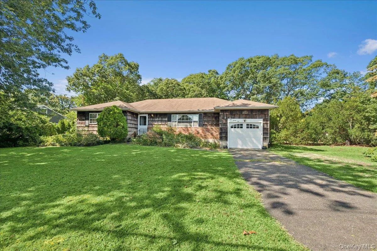 Ranch-style residence featuring wood shingle siding, an attached garage with a white panel door, a paved driveway, and a spacious front lawn Ranch-style residence featuring wood shingle siding, an attached garage with a white panel door, a paved driveway, and a spacious front lawn