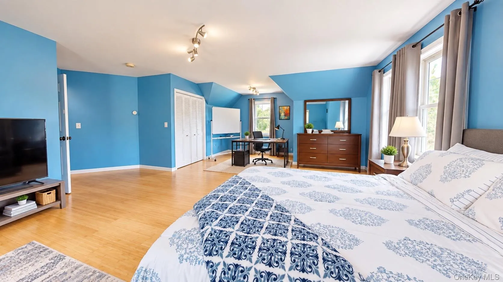 Spacious room with wood-finish flooring, multiple windows, track lighting, and a built-in closet with bi-fold doors Spacious room with wood-finish flooring, multiple windows, track lighting, and a built-in closet with bi-fold doors
