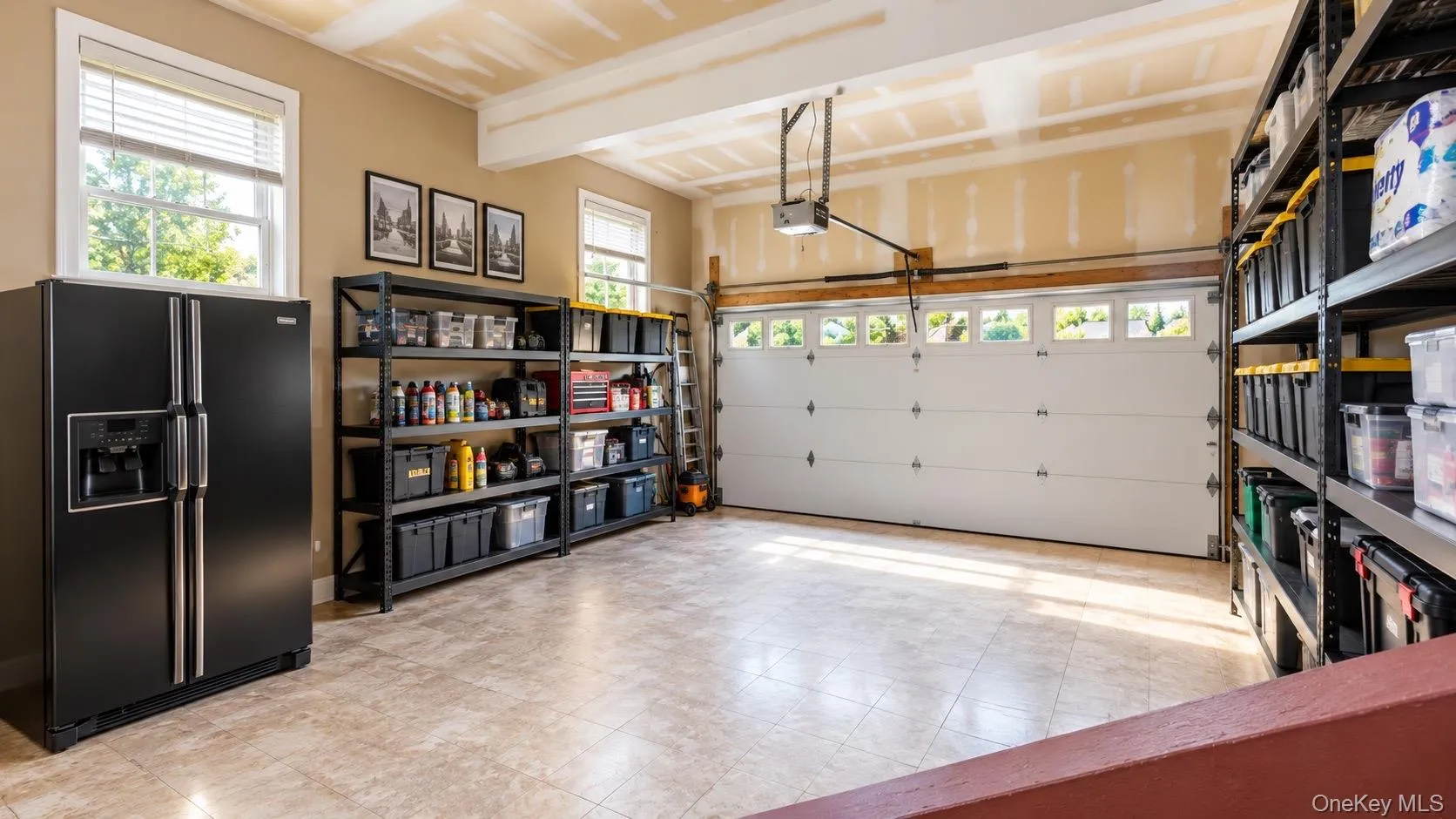 Attached two-car garage featuring a garage door with transom windows, two large windows, exposed ceiling beams, and light-toned tile flooring Attached two-car garage featuring a garage door with transom windows, two large windows, exposed ceiling beams, and light-toned tile flooring