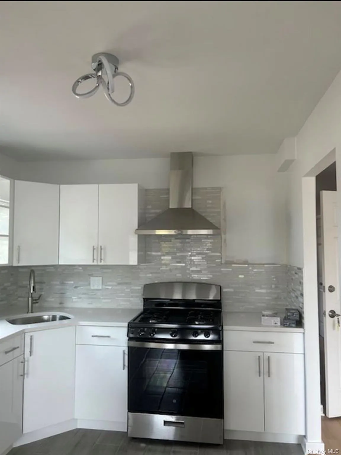 Sleek kitchen featuring white cabinetry, light countertops, a stainless steel gas range, a coordinating range hood, and a mosaic tile backsplash Sleek kitchen featuring white cabinetry, light countertops, a stainless steel gas range, a coordinating range hood, and a mosaic tile backsplash