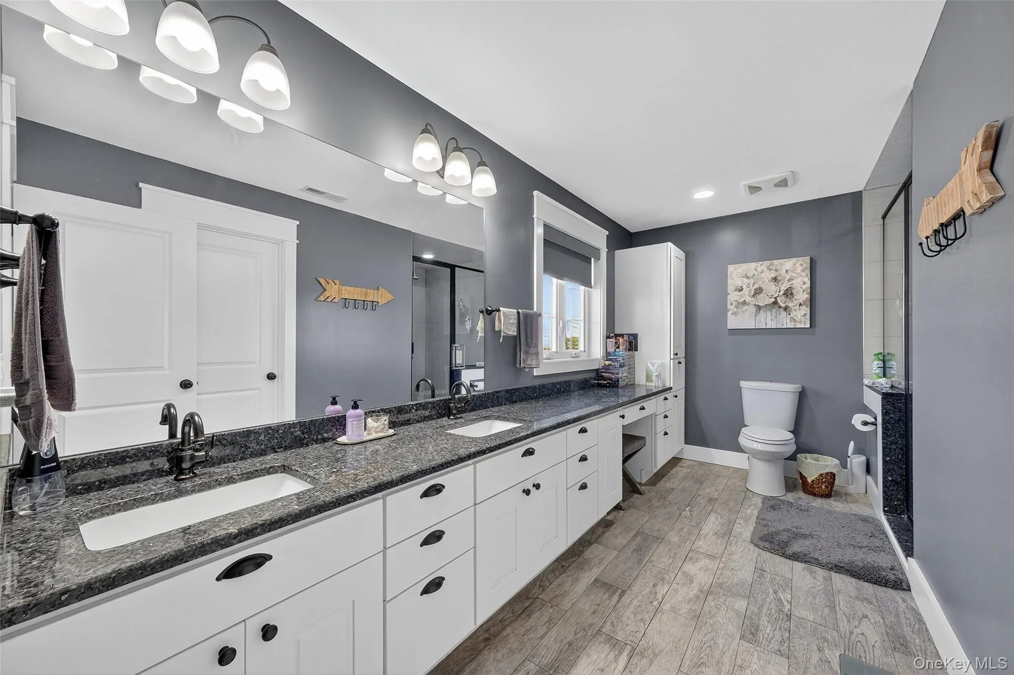 Dual vanities with undermount sinks, wood-finish flooring, a built-in linen cabinet, a window with roller shade, and a glass-enclosed shower Dual vanities with undermount sinks, wood-finish flooring, a built-in linen cabinet, a window with roller shade, and a glass-enclosed shower