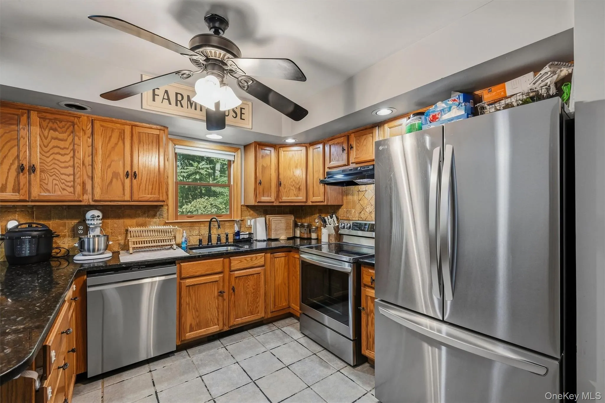 Kitchen featuring wood cabinetry, dark countertops, stainless steel appliances, tile flooring, and a ceiling fan with integrated lighting Kitchen featuring wood cabinetry, dark countertops, stainless steel appliances, tile flooring, and a ceiling fan with integrated lighting
