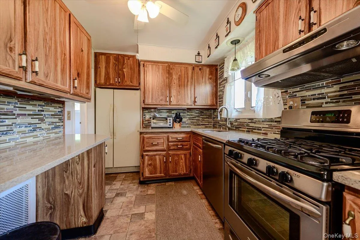 Kitchen featuring wood-finish cabinetry, stainless steel range and hood, mosaic tile backsplash, light-toned countertops, and ceiling fan Kitchen featuring wood-finish cabinetry, stainless steel range and hood, mosaic tile backsplash, light-toned countertops, and ceiling fan