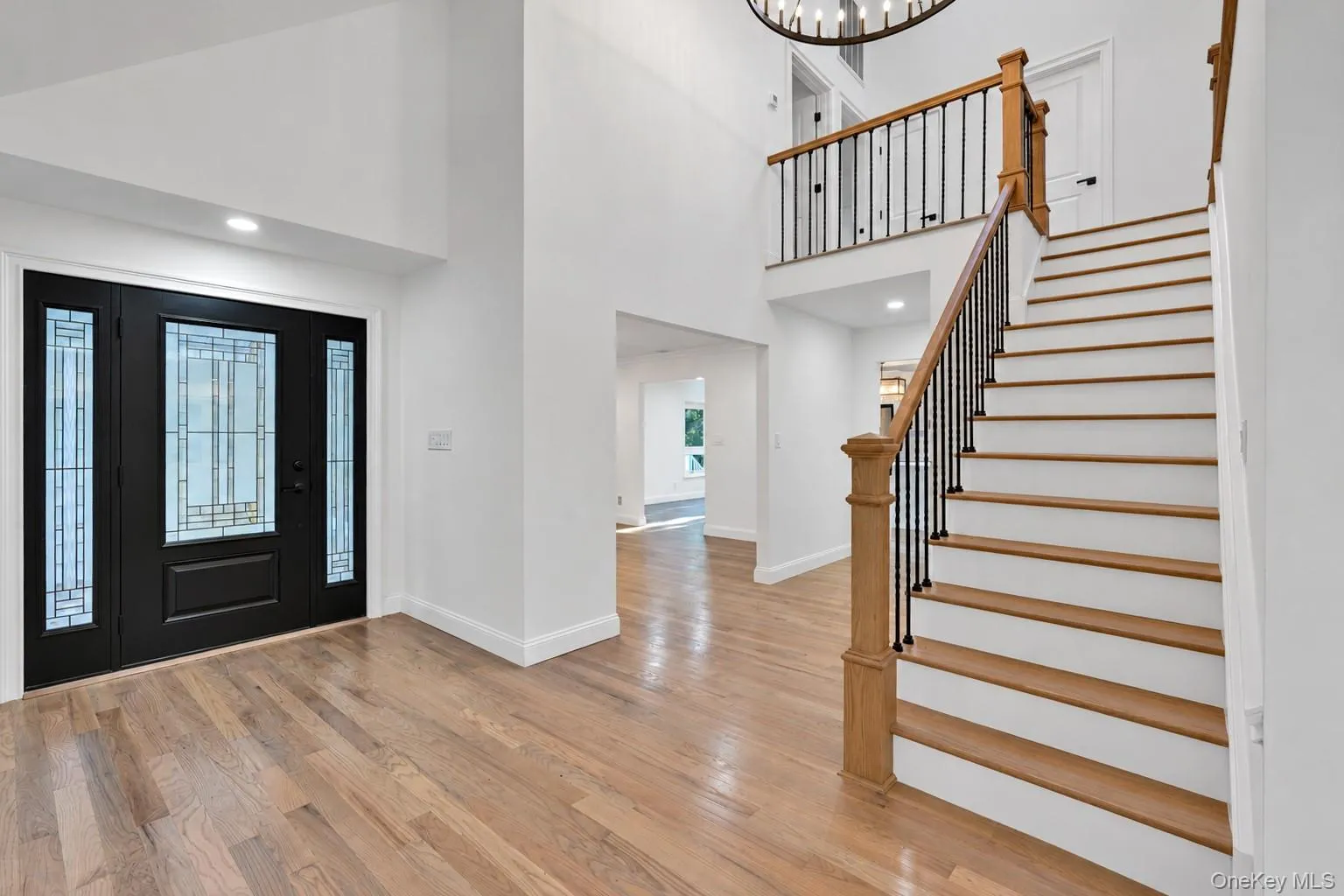 Two-story foyer featuring wide plank wood-finish flooring and a black front entry door with leaded glass sidelights Two-story foyer featuring wide plank wood-finish flooring and a black front entry door with leaded glass sidelights