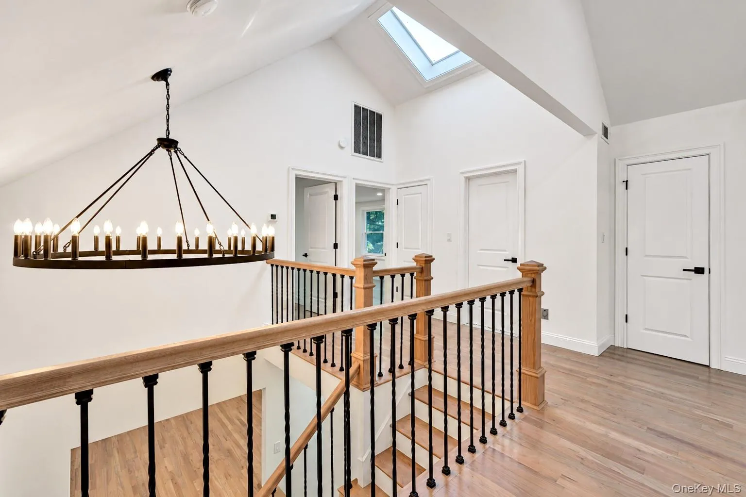 Vaulted ceiling with skylight, wood-finish flooring, wrought iron chandelier, wood handrail with black balusters, and white paneled doors with matte black hardware Vaulted ceiling with skylight, wood-finish flooring, wrought iron chandelier, wood handrail with black balusters, and white paneled doors with matte black hardware