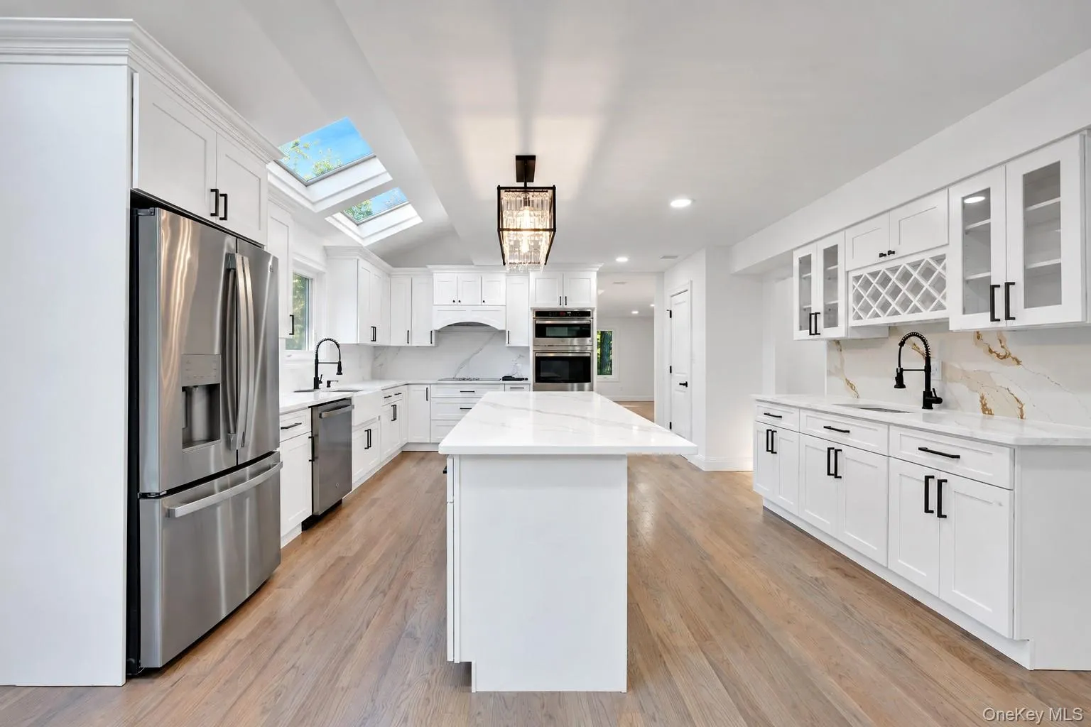 Modern kitchen featuring white cabinetry, a large center island with a stone countertop, stainless steel appliances, wood-finish flooring, and dual skylights Modern kitchen featuring white cabinetry, a large center island with a stone countertop, stainless steel appliances, wood-finish flooring, and dual skylights
