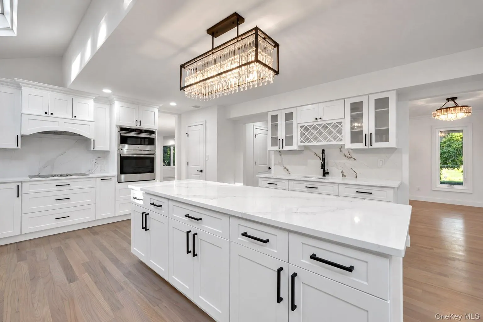 White shaker cabinetry with black hardware, white countertops and backsplash, stainless steel double wall oven, and light wood-finish flooring White shaker cabinetry with black hardware, white countertops and backsplash, stainless steel double wall oven, and light wood-finish flooring