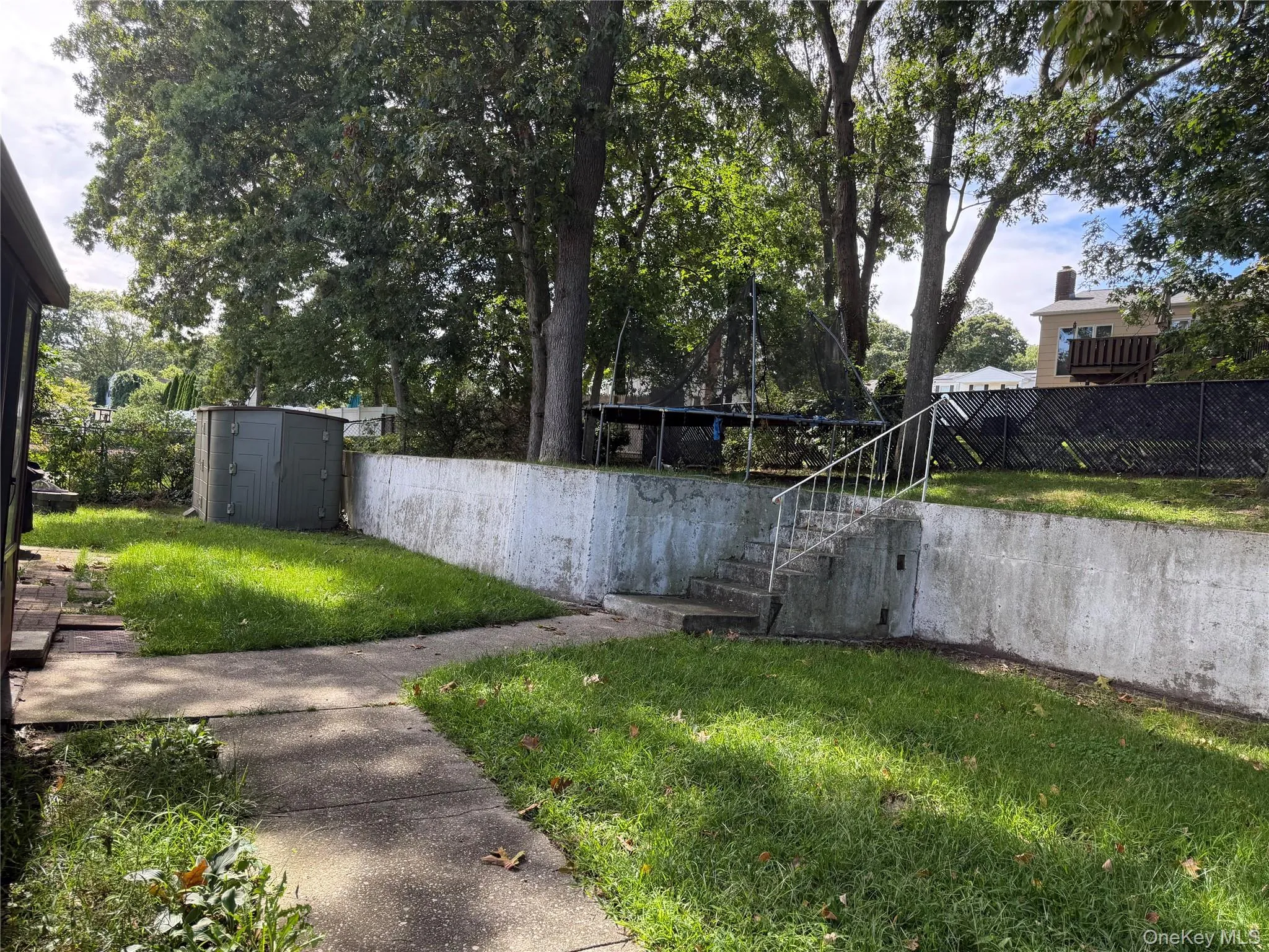 Manicured lawn with mature trees, concrete pathway, retaining wall, and exterior storage shed Manicured lawn with mature trees, concrete pathway, retaining wall, and exterior storage shed