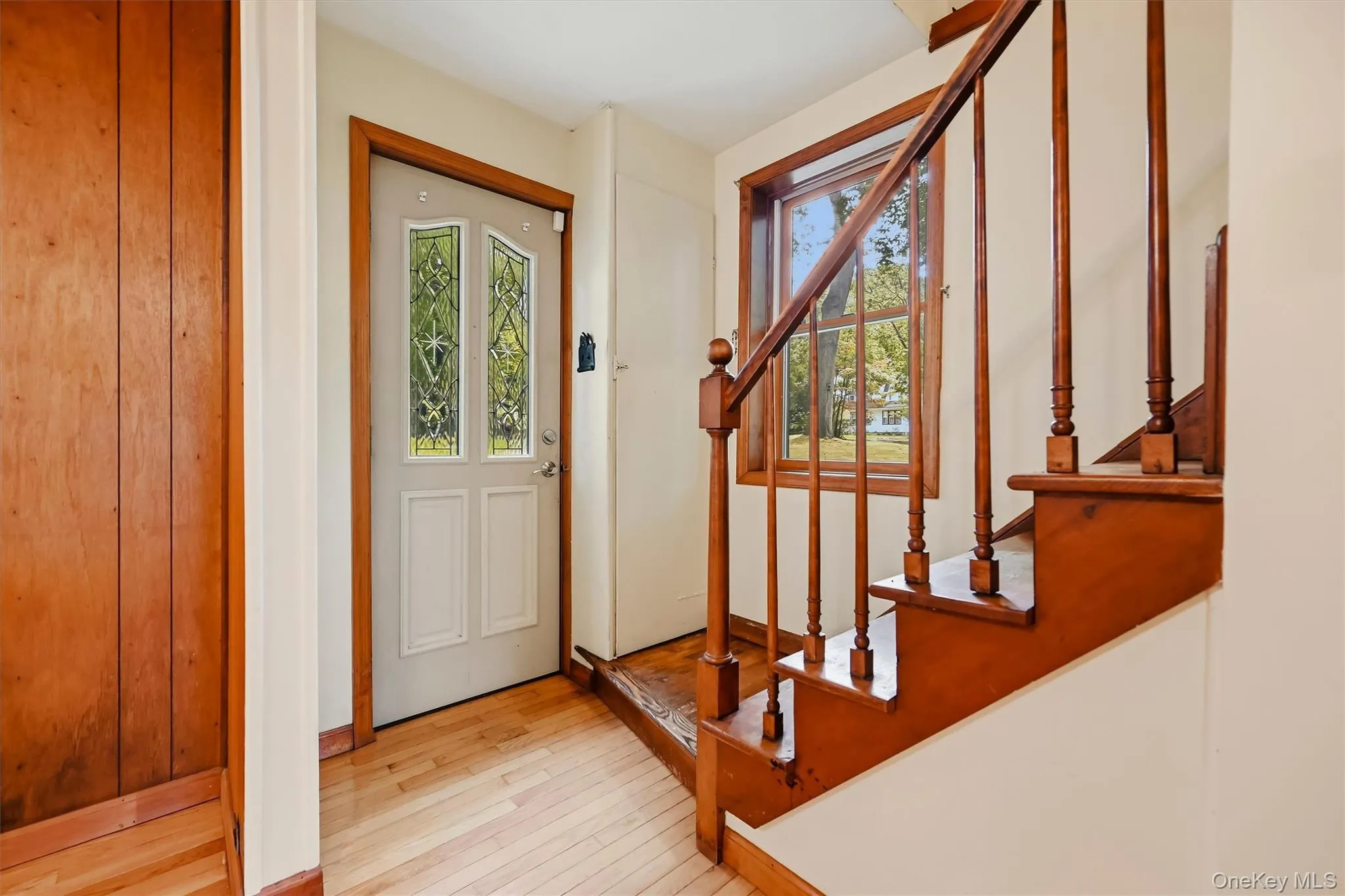 Hardwood flooring, wood-paneled wall, white entry door with decorative glass inserts, wood staircase with balusters, and a wood-framed window Hardwood flooring, wood-paneled wall, white entry door with decorative glass inserts, wood staircase with balusters, and a wood-framed window