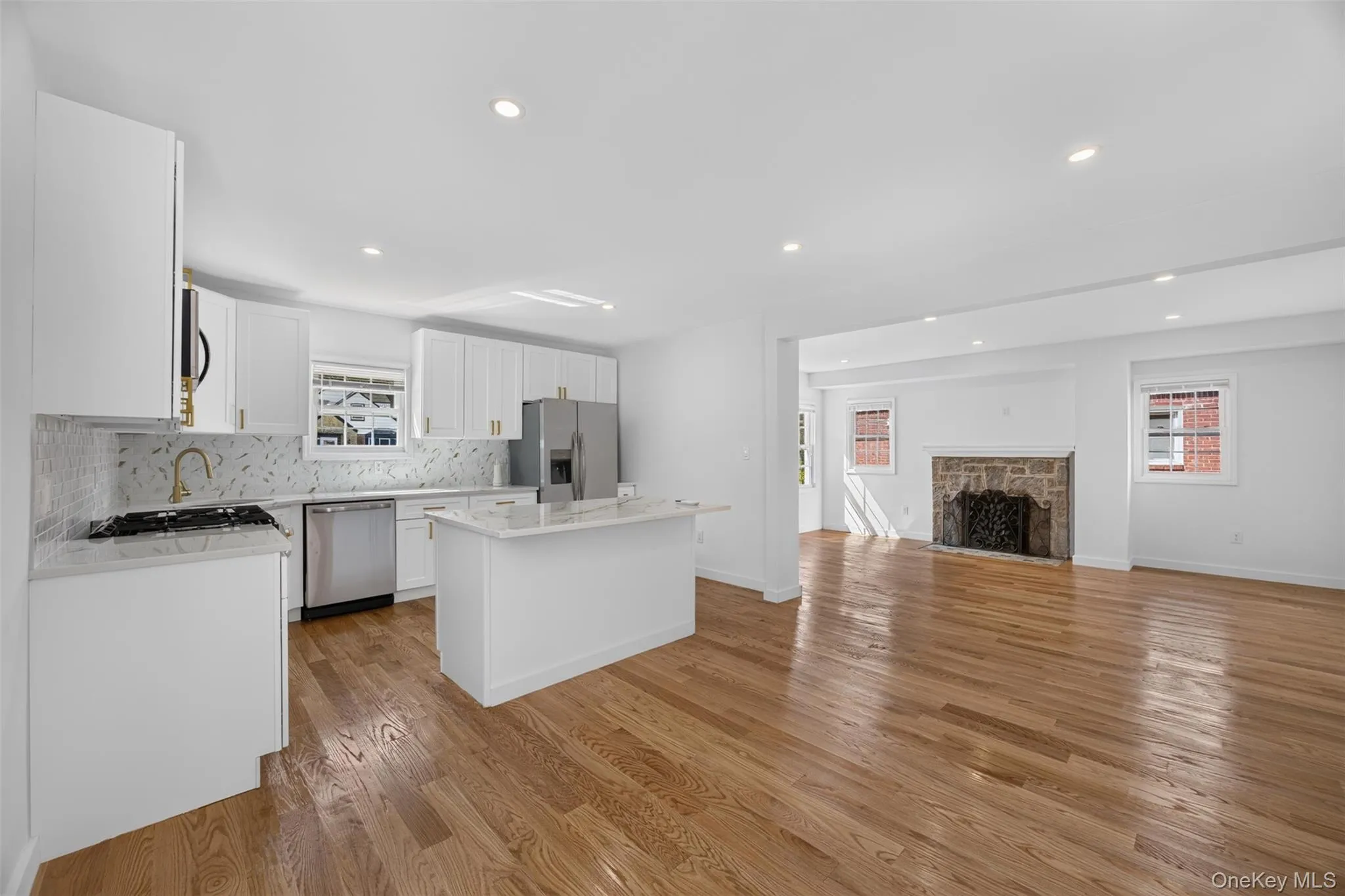 Open-concept living space featuring wood-finish flooring, a stone fireplace, recessed lighting, white cabinetry, and stainless steel appliances Open-concept living space featuring wood-finish flooring, a stone fireplace, recessed lighting, white cabinetry, and stainless steel appliances