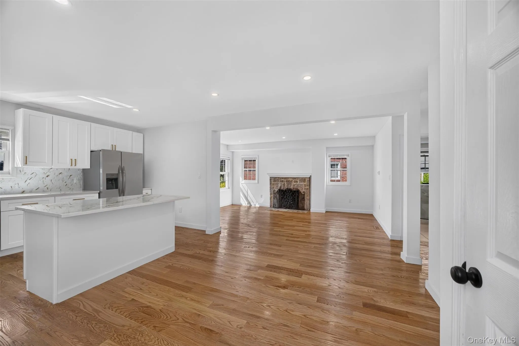 Refinished hardwood flooring, recessed lighting, white kitchen cabinetry with a stainless steel refrigerator, stone fireplace, and a kitchen island with a light-colored countertop Refinished hardwood flooring, recessed lighting, white kitchen cabinetry with a stainless steel refrigerator, stone fireplace, and a kitchen island with a light-colored countertop