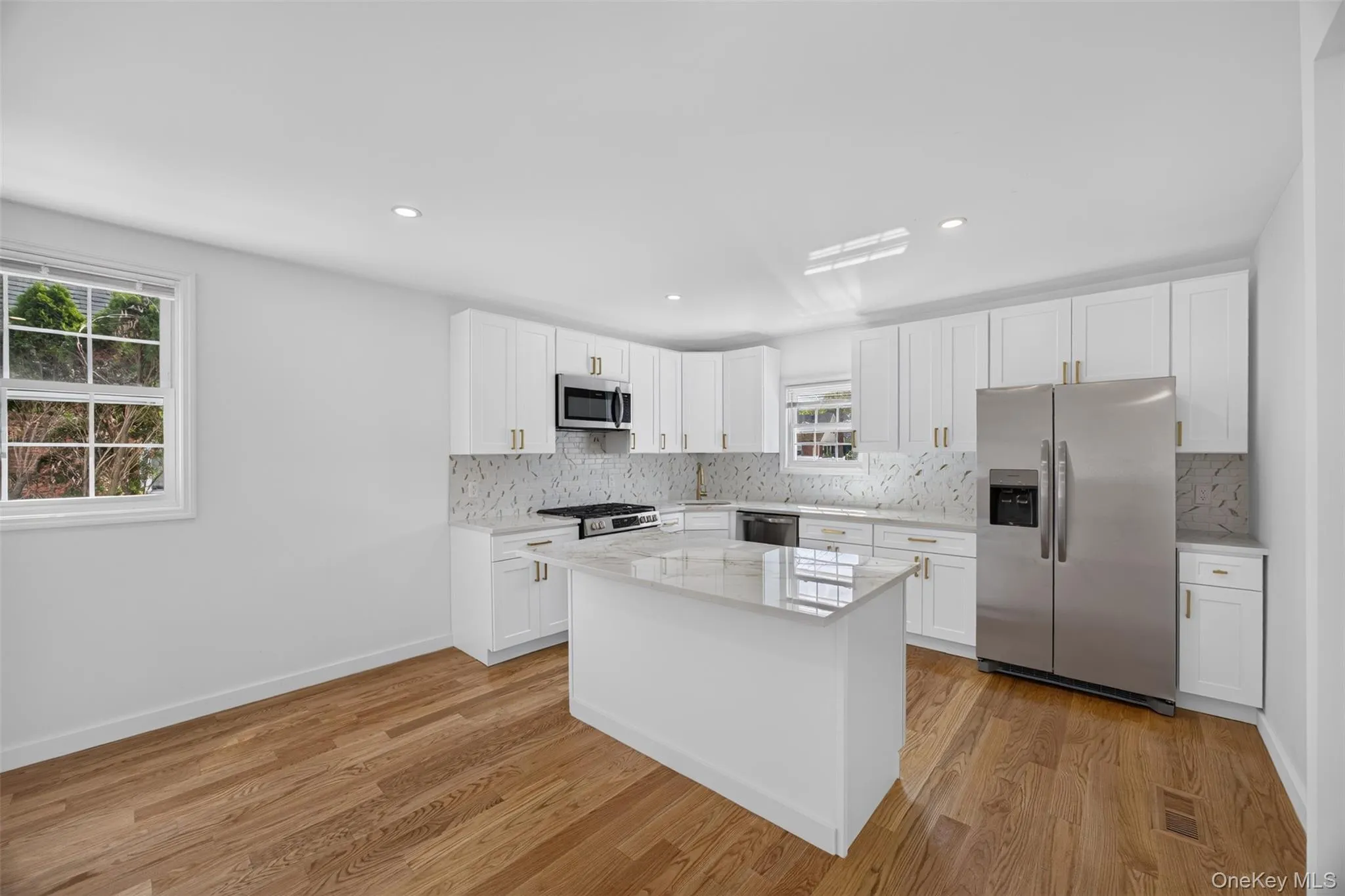 Kitchen featuring wood-finish flooring, white cabinetry with gold-tone hardware, a center island with a stone countertop, stainless steel appliances, and recessed lighting Kitchen featuring wood-finish flooring, white cabinetry with gold-tone hardware, a center island with a stone countertop, stainless steel appliances, and recessed lighting