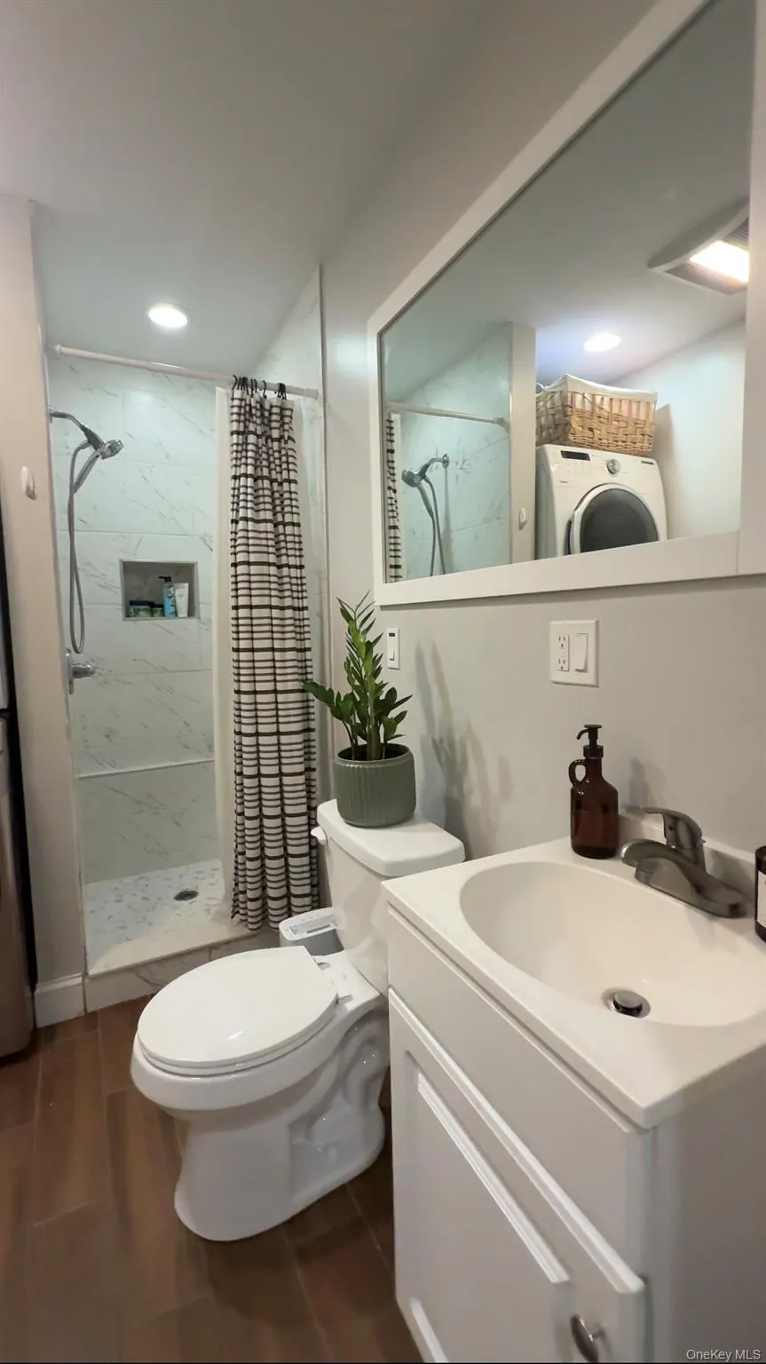 Cottage full bathroom with wood-finish flooring, stacked laundry appliances, walk-in shower with glass enclosure, recessed lighting, and a vanity with an integrated sink Cottage full bathroom with wood-finish flooring, stacked laundry appliances, walk-in shower with glass enclosure, recessed lighting, and a vanity with an integrated sink