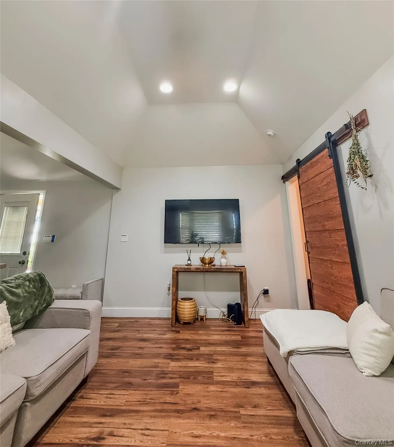 Cottage living room with vaulted ceiling with recessed lighting, wood-finish flooring, and a sliding wood barn door Cottage living room with vaulted ceiling with recessed lighting, wood-finish flooring, and a sliding wood barn door