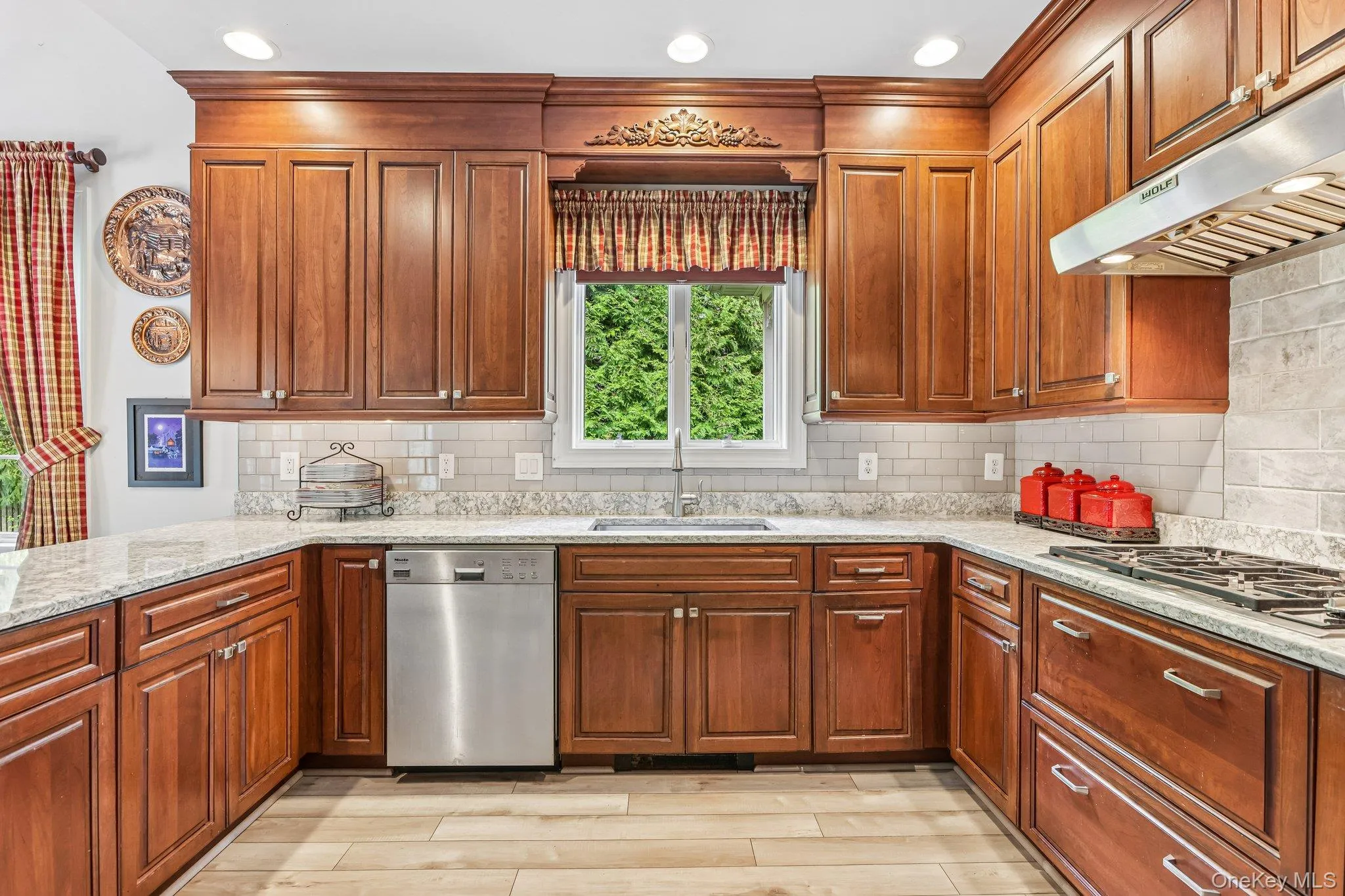 Cherry wood cabinetry, light-toned countertops, subway tile backsplash, stainless steel dishwasher, and a gas cooktop Cherry wood cabinetry, light-toned countertops, subway tile backsplash, stainless steel dishwasher, and a gas cooktop