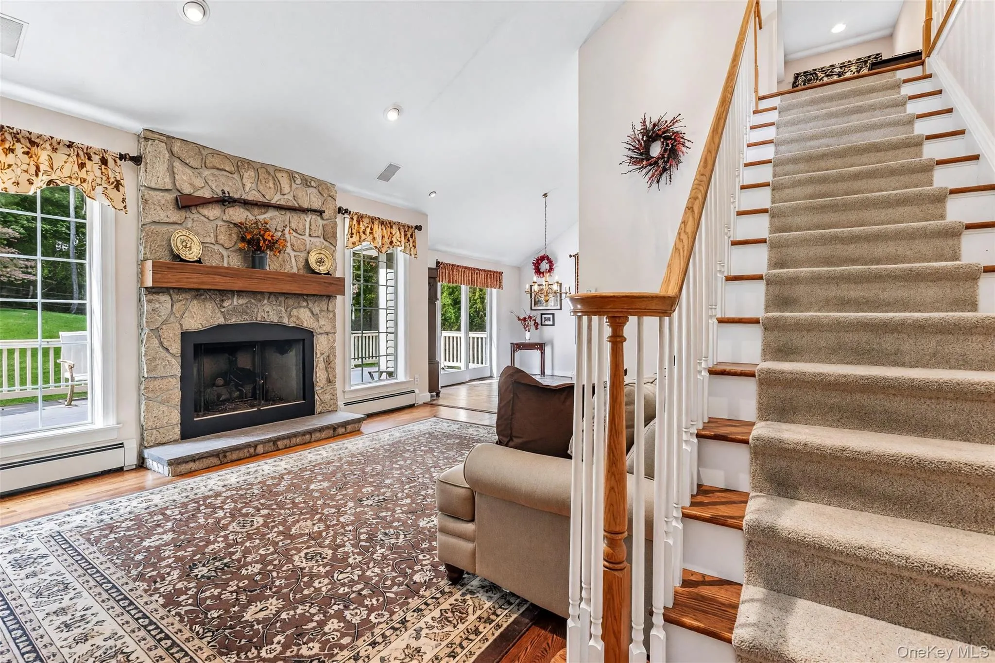 Stone-clad fireplace with a wood mantle, hardwood flooring, and a carpeted staircase with a wood handrail Stone-clad fireplace with a wood mantle, hardwood flooring, and a carpeted staircase with a wood handrail