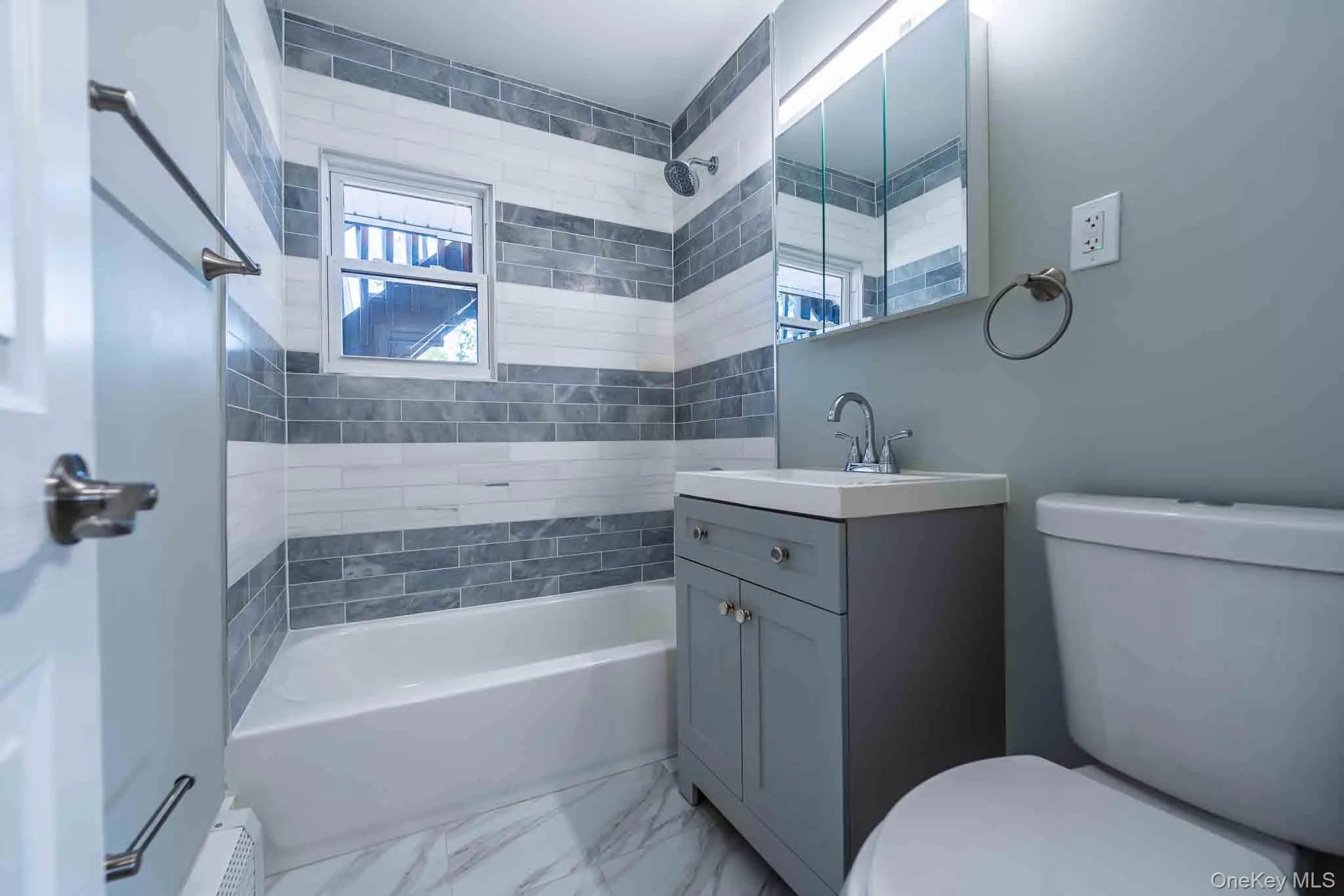 New Contemporary bathroom featuring a white bathtub, gray and white striped tile surround, single vanity with integrated sink, chrome fixtures, and a mirrored medicine cabinet with integrated lighting New Contemporary bathroom featuring a white bathtub, gray and white striped tile surround, single vanity with integrated sink, chrome fixtures, and a mirrored medicine cabinet with integrated lighting