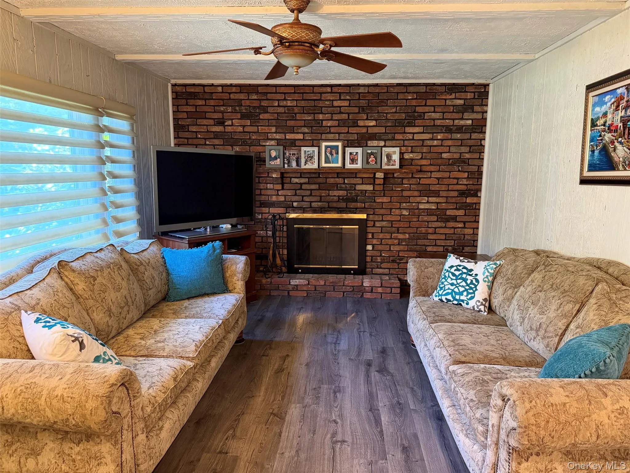 Brick accent wall with fireplace, wood-finish flooring, ceiling fan, and window with horizontal blinds Brick accent wall with fireplace, wood-finish flooring, ceiling fan, and window with horizontal blinds