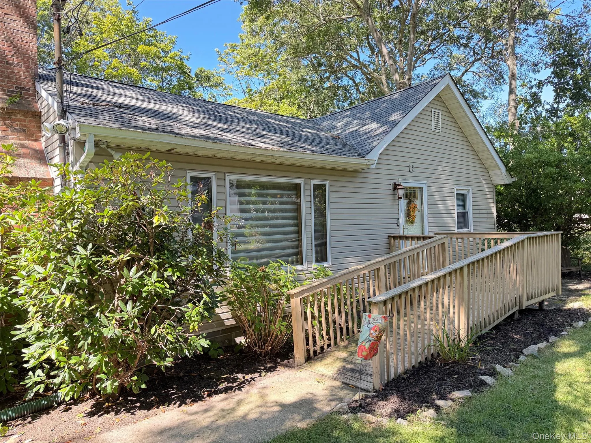 Gable roofline with light-colored siding, extensive wood ramp, prominent window with blinds, and lush landscaping Gable roofline with light-colored siding, extensive wood ramp, prominent window with blinds, and lush landscaping
