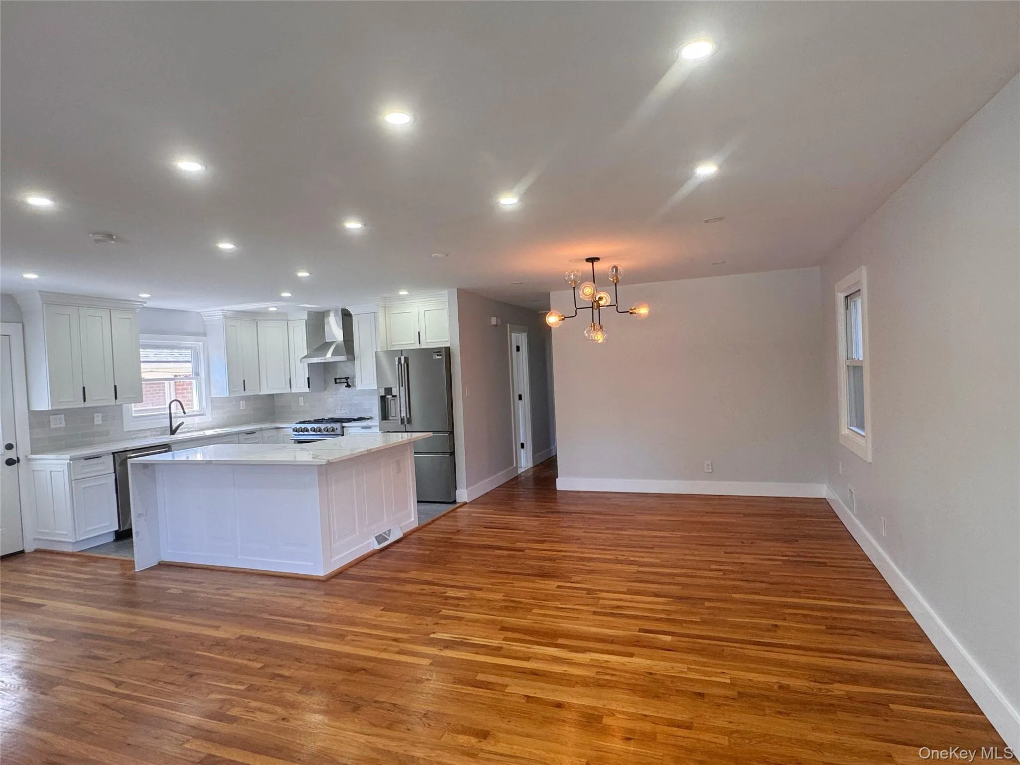 Refinished hardwood flooring throughout the main living area, a kitchen featuring white cabinetry, light countertops, and stainless steel appliances, recessed ceiling lighting, and a contemporary ceiling fixture Refinished hardwood flooring throughout the main living area, a kitchen featuring white cabinetry, light countertops, and stainless steel appliances, recessed ceiling lighting, and a contemporary ceiling fixture