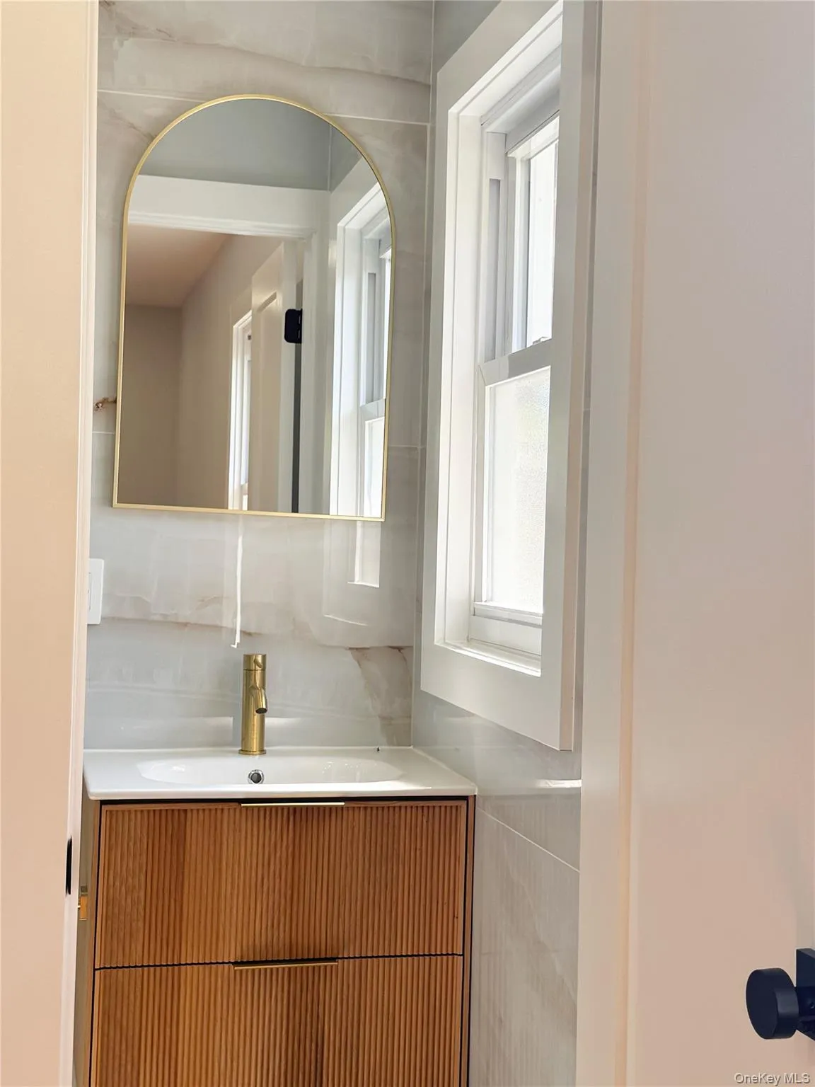 Bathroom featuring a wood-finish vanity with an integrated sink, gold-tone faucet, arched mirror, and tiled walls Bathroom featuring a wood-finish vanity with an integrated sink, gold-tone faucet, arched mirror, and tiled walls