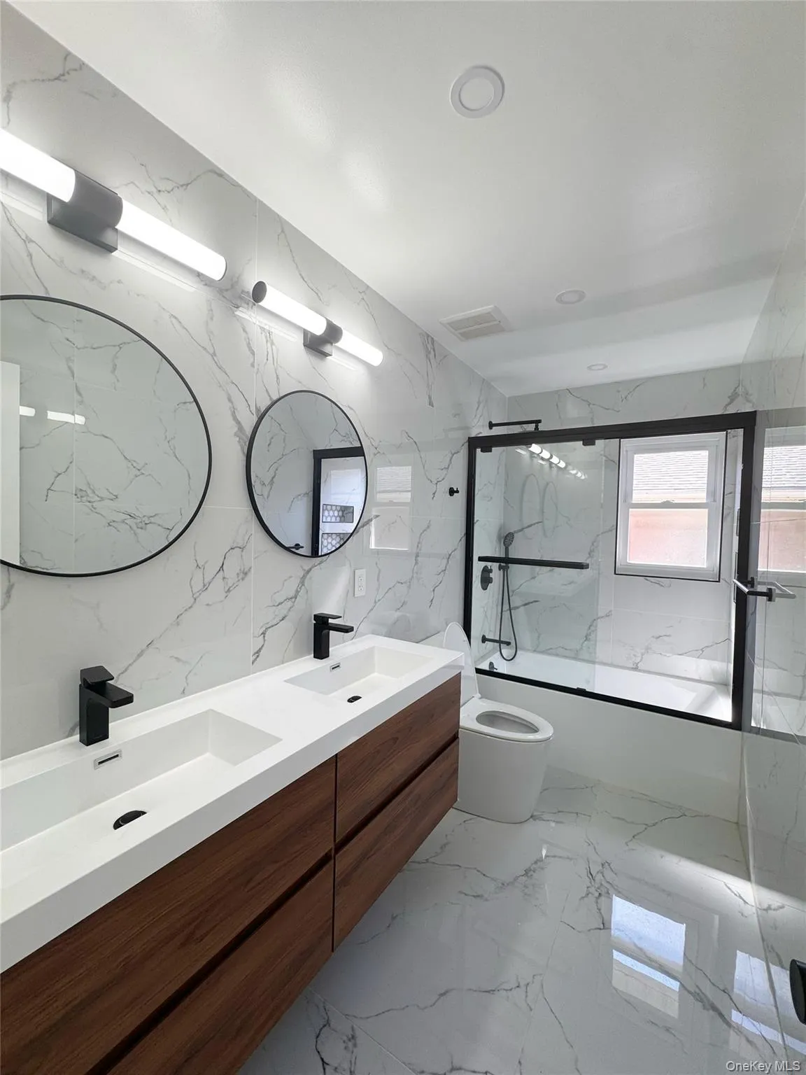 Dual vessel sinks with matte black faucets, wood-finish floating vanity, round framed mirrors, large format marble-look wall and floor tile, and a tub with a black-framed glass enclosure Dual vessel sinks with matte black faucets, wood-finish floating vanity, round framed mirrors, large format marble-look wall and floor tile, and a tub with a black-framed glass enclosure