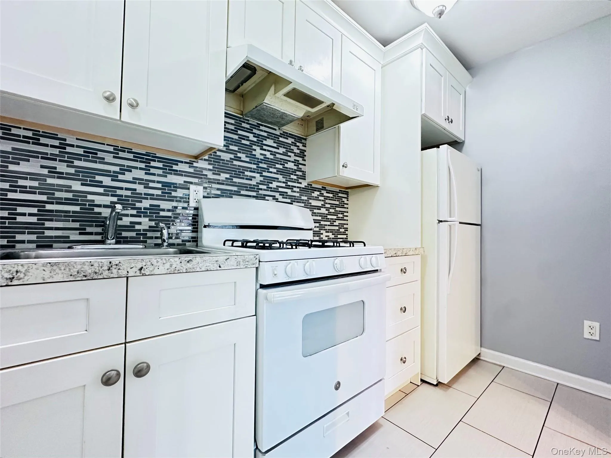 White shaker-style cabinetry, mosaic tile backsplash, speckled countertops, gas range, spacious refrigerator and tile flooring White shaker-style cabinetry, mosaic tile backsplash, speckled countertops, gas range, spacious refrigerator and tile flooring