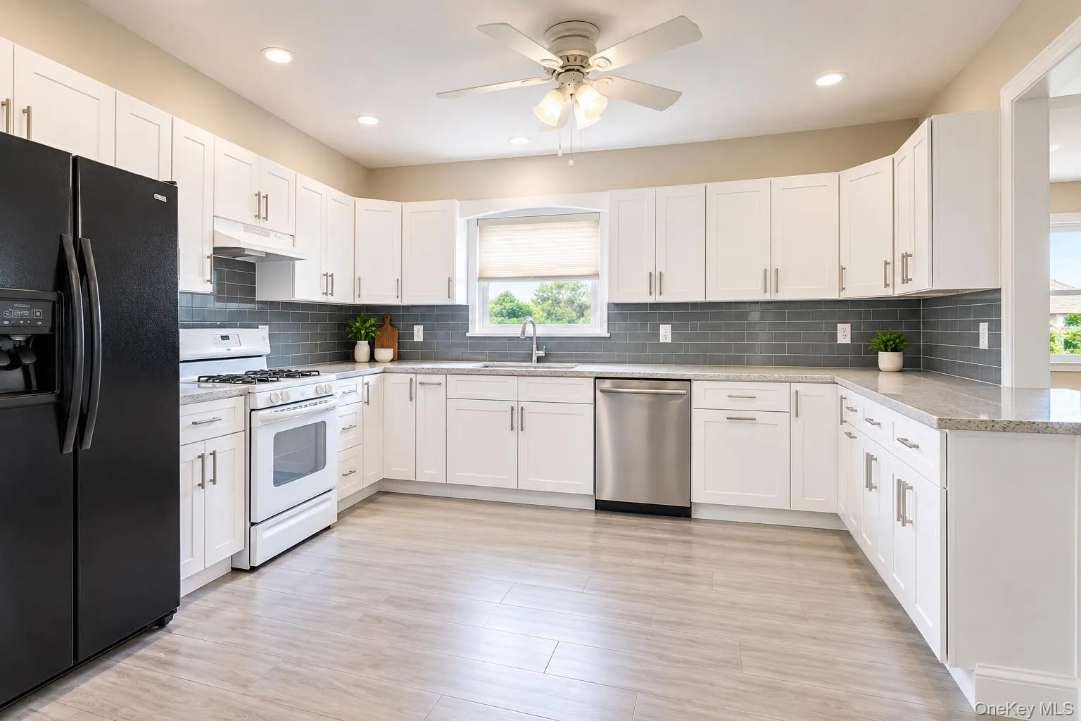 Bright kitchen with white cabinetry, light countertops, gray subway tile backsplash, stainless steel dishwasher, and wood-finish flooring Bright kitchen with white cabinetry, light countertops, gray subway tile backsplash, stainless steel dishwasher, and wood-finish flooring