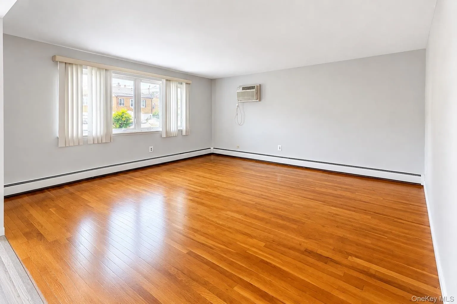 Hardwood flooring, light gray walls, large window with vertical blinds, and a wall-mounted air conditioning unit Hardwood flooring, light gray walls, large window with vertical blinds, and a wall-mounted air conditioning unit