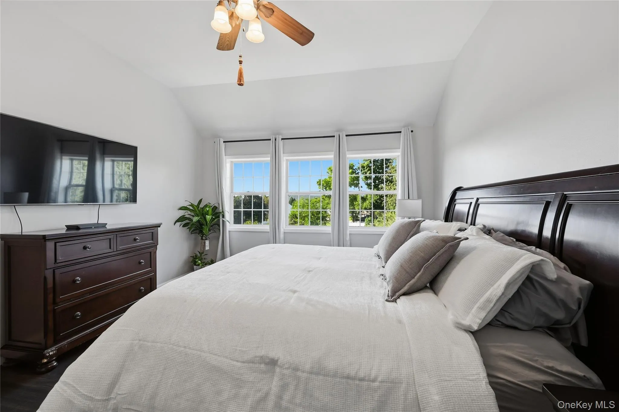 Vaulted ceiling, wood-finish flooring, ceiling fan with light kit, three large windows, and dark wood headboard Vaulted ceiling, wood-finish flooring, ceiling fan with light kit, three large windows, and dark wood headboard