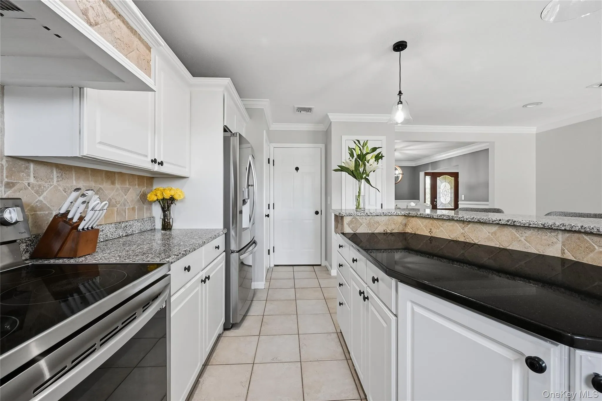 White cabinetry with black hardware, stainless steel appliances, stone tile backsplash, granite countertops, and tile flooring White cabinetry with black hardware, stainless steel appliances, stone tile backsplash, granite countertops, and tile flooring