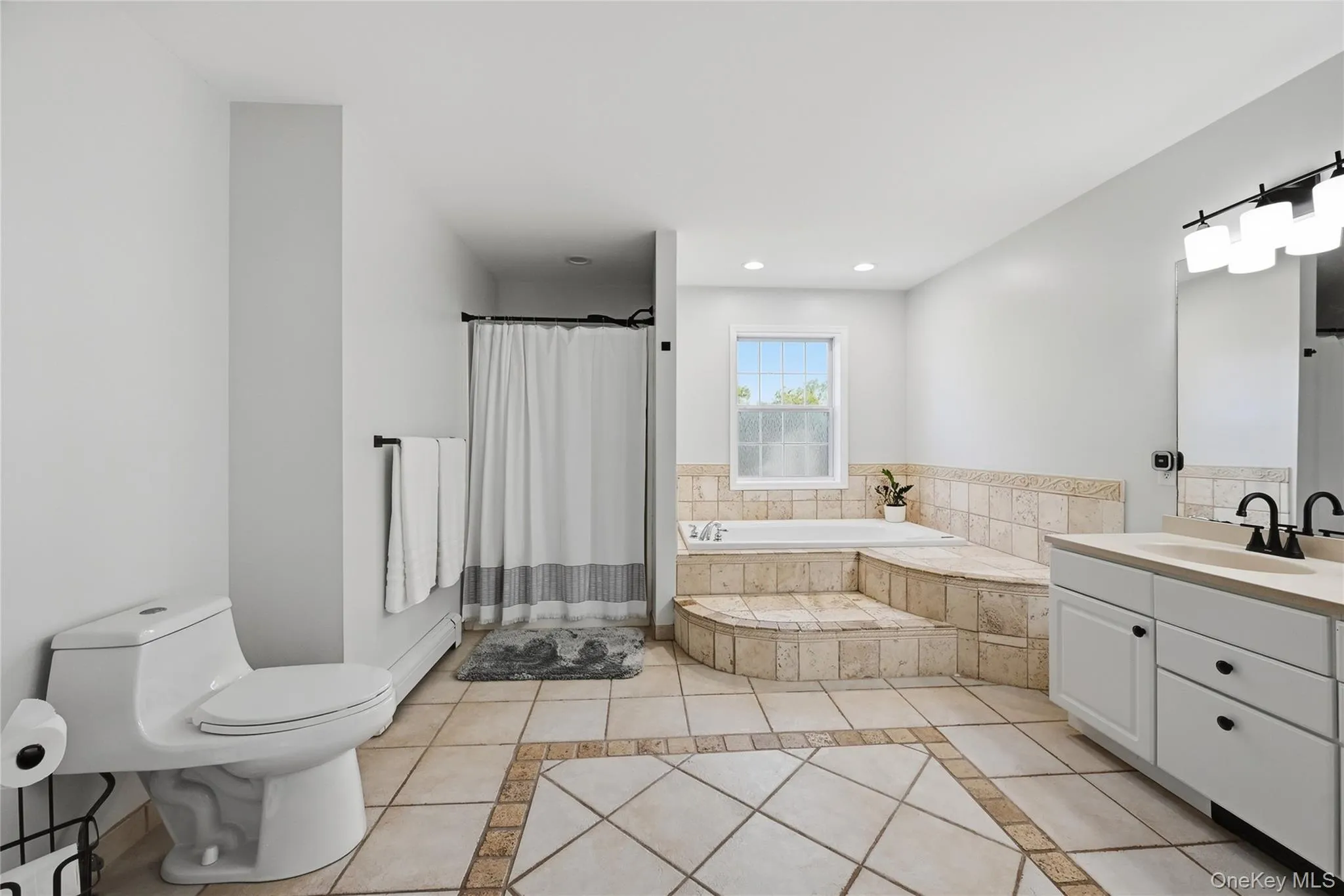 Soaking tub with tiled surround, white vanity with dual sinks, tiled flooring with accent border, and a separate shower enclosure Soaking tub with tiled surround, white vanity with dual sinks, tiled flooring with accent border, and a separate shower enclosure