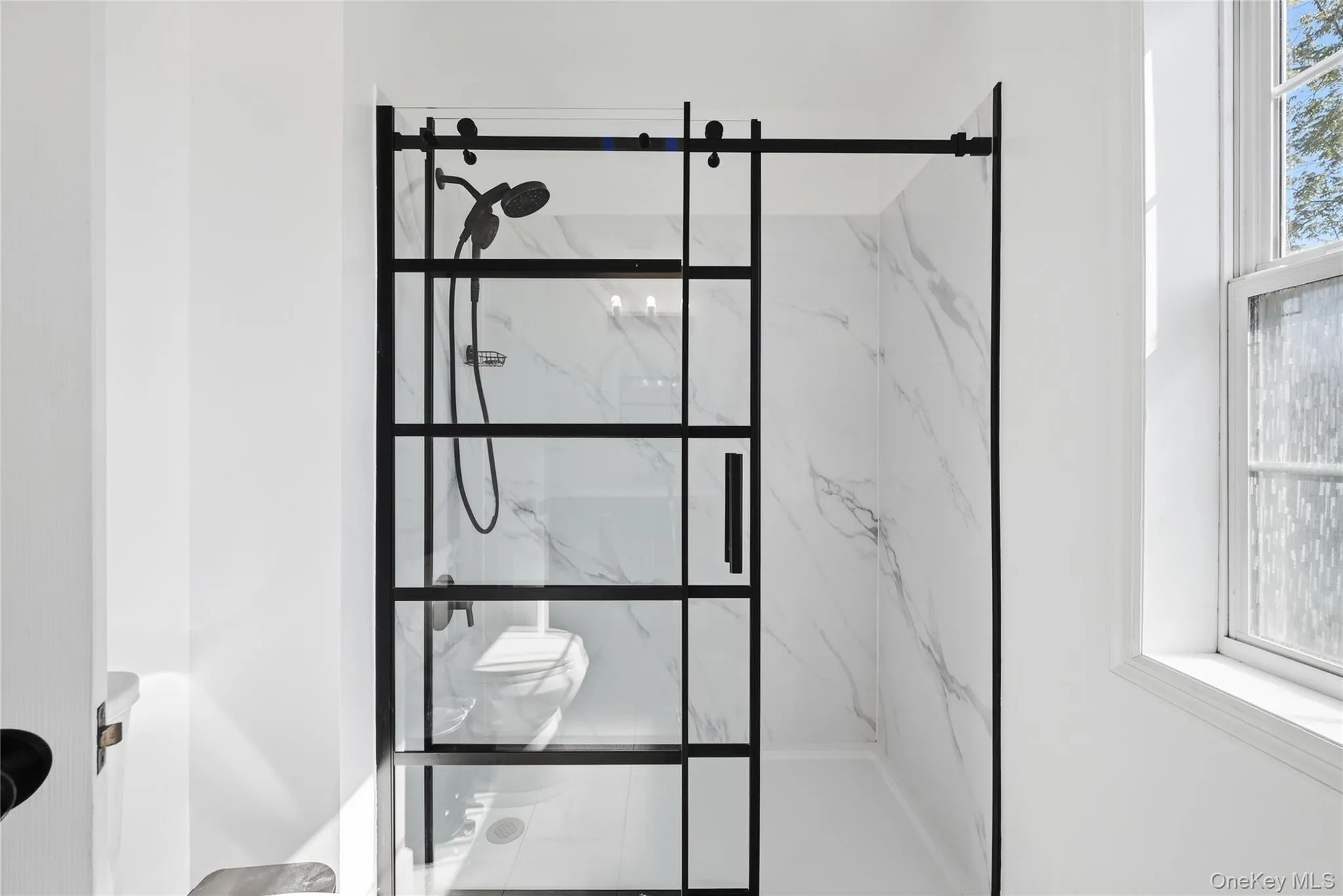 Walk-in shower featuring a sliding glass door with black framing, matte black shower fixtures, and white tile with gray veining Walk-in shower featuring a sliding glass door with black framing, matte black shower fixtures, and white tile with gray veining