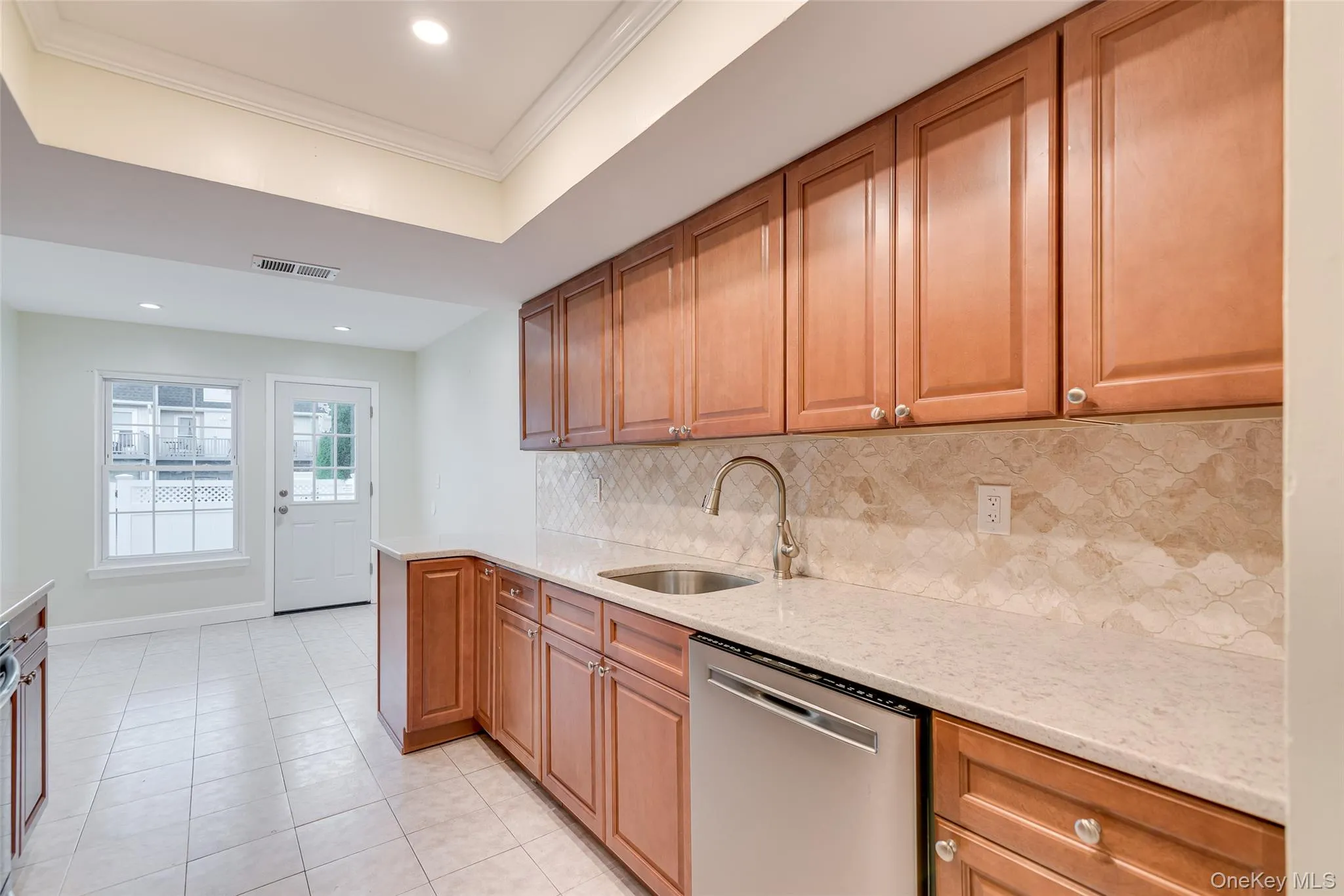 Wood-finish cabinetry, light-toned countertops, tile backsplash, stainless steel dishwasher, and recessed lighting Wood-finish cabinetry, light-toned countertops, tile backsplash, stainless steel dishwasher, and recessed lighting
