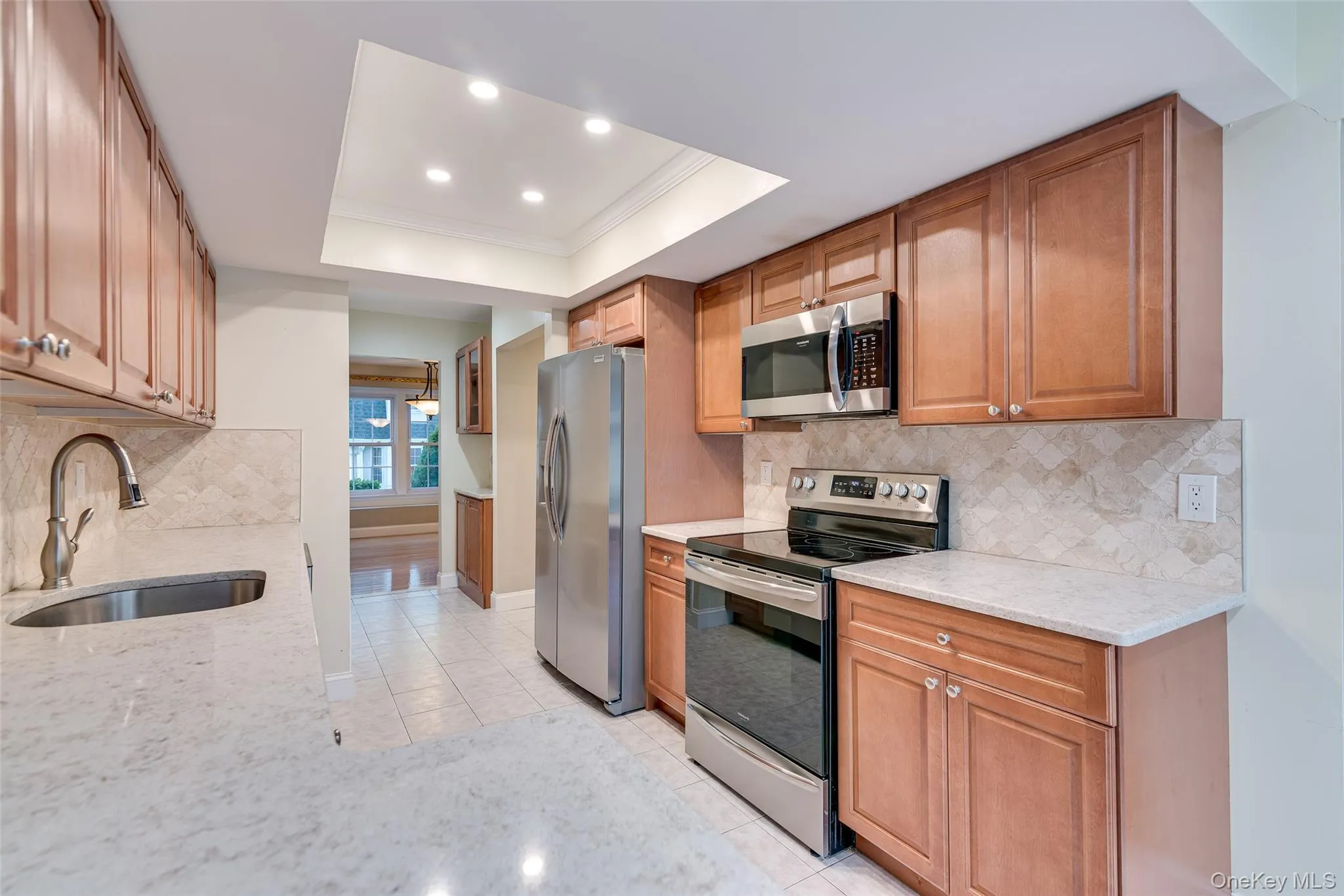 Kitchen featuring wood cabinetry, stainless steel appliances, light-toned countertops, tile flooring, and a patterned tile backsplash Kitchen featuring wood cabinetry, stainless steel appliances, light-toned countertops, tile flooring, and a patterned tile backsplash