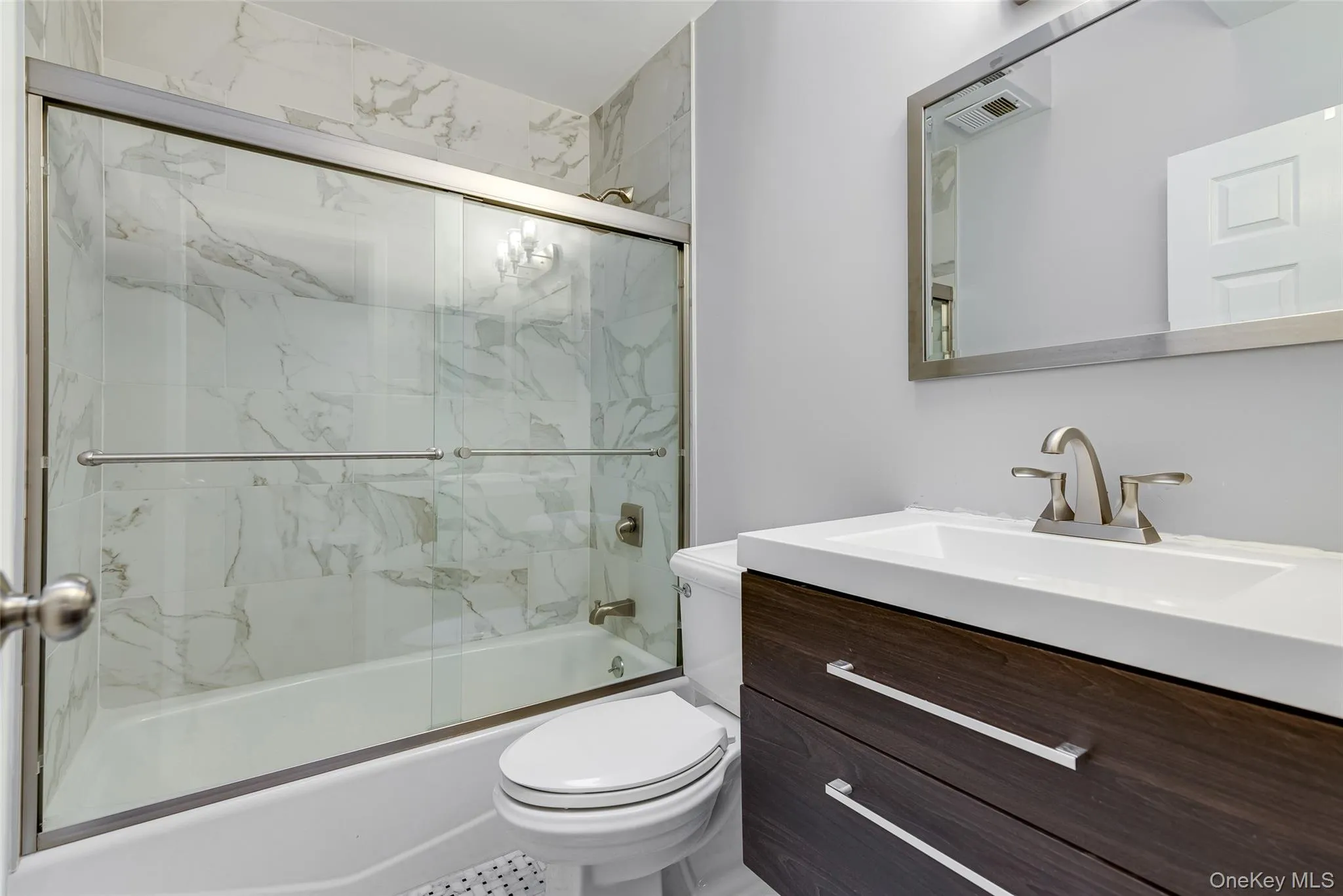 Tub and shower combination with sliding glass doors, marble-look tiled surround, and a vanity with a white countertop and dark wood-finish cabinetry Tub and shower combination with sliding glass doors, marble-look tiled surround, and a vanity with a white countertop and dark wood-finish cabinetry