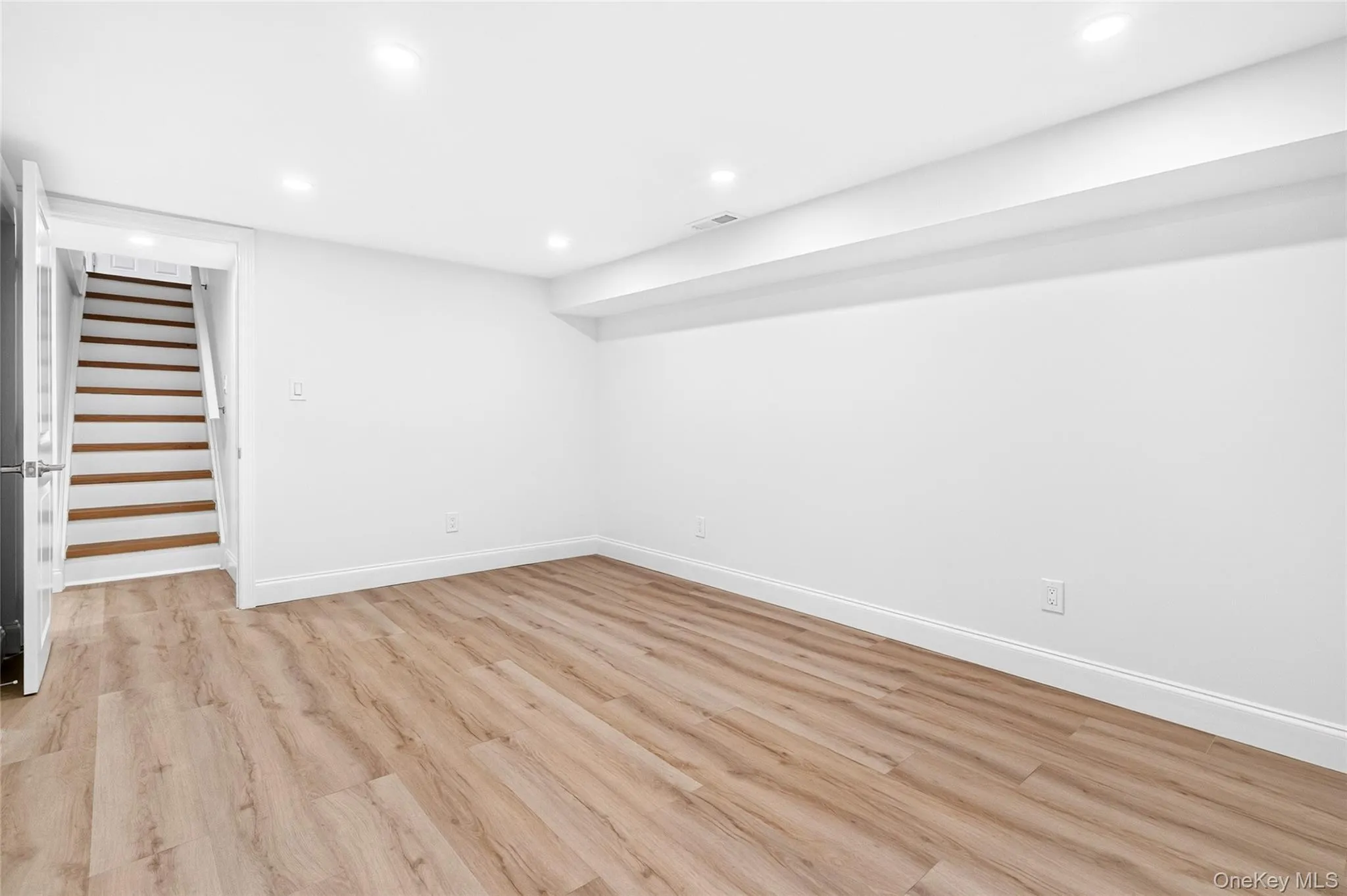 Recessed lighting illuminating the room, wood-finish flooring, white painted walls, and a staircase with wood treads Recessed lighting illuminating the room, wood-finish flooring, white painted walls, and a staircase with wood treads