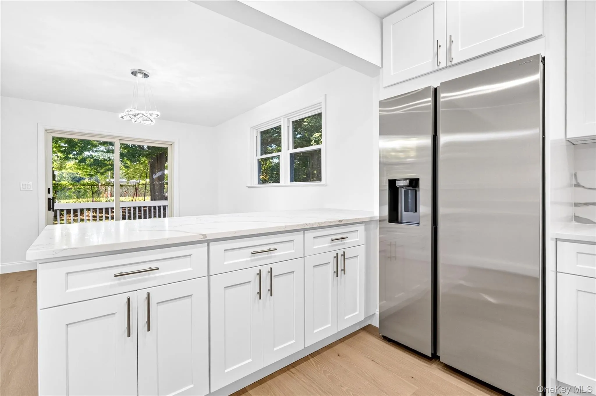White shaker cabinetry, light-toned wood-finish flooring, stainless steel refrigerator, white countertops, and a contemporary ceiling fixture White shaker cabinetry, light-toned wood-finish flooring, stainless steel refrigerator, white countertops, and a contemporary ceiling fixture