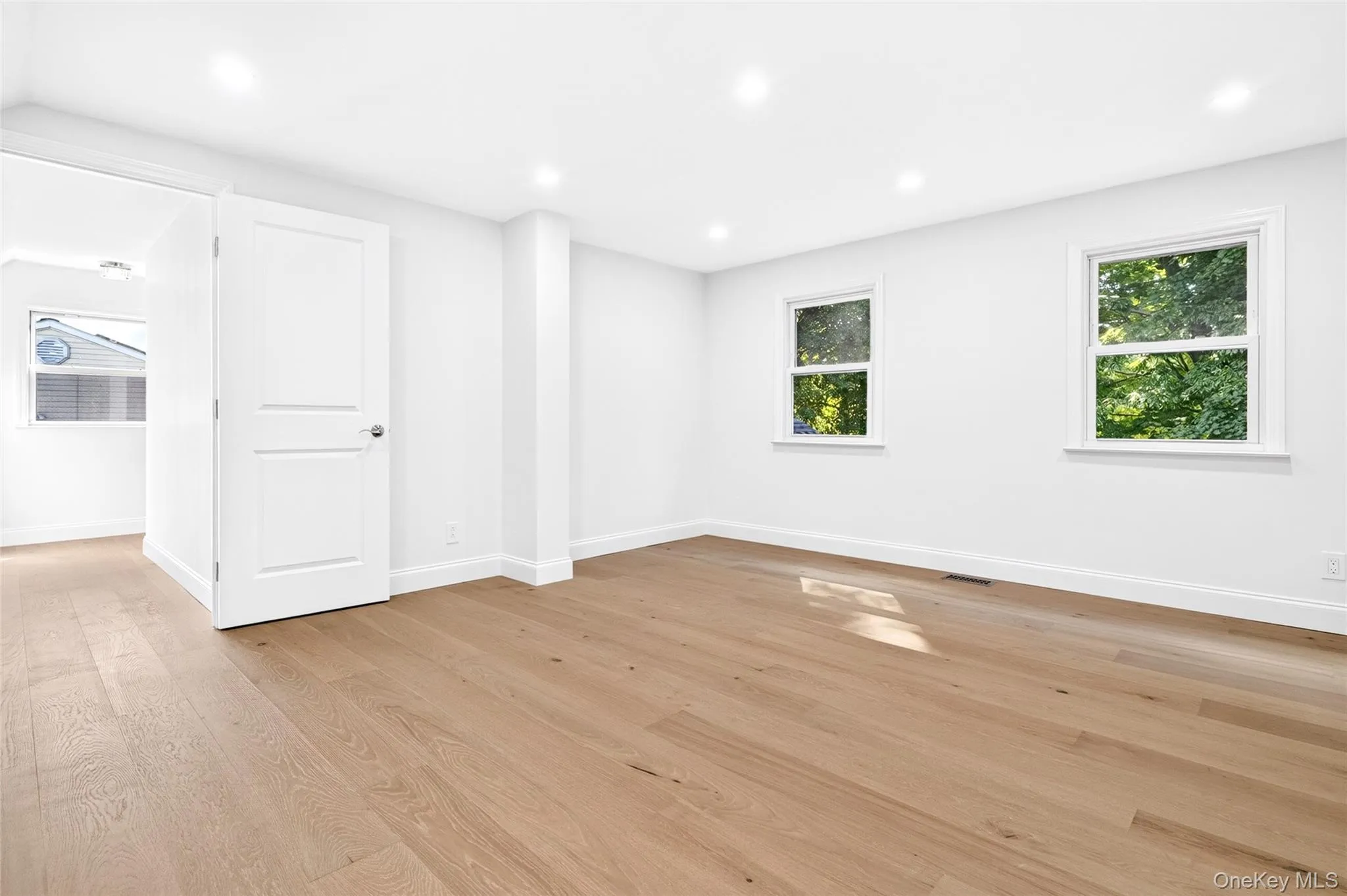 Recessed lighting and two windows illuminate this room with wood-finish flooring and a white paneled door Recessed lighting and two windows illuminate this room with wood-finish flooring and a white paneled door