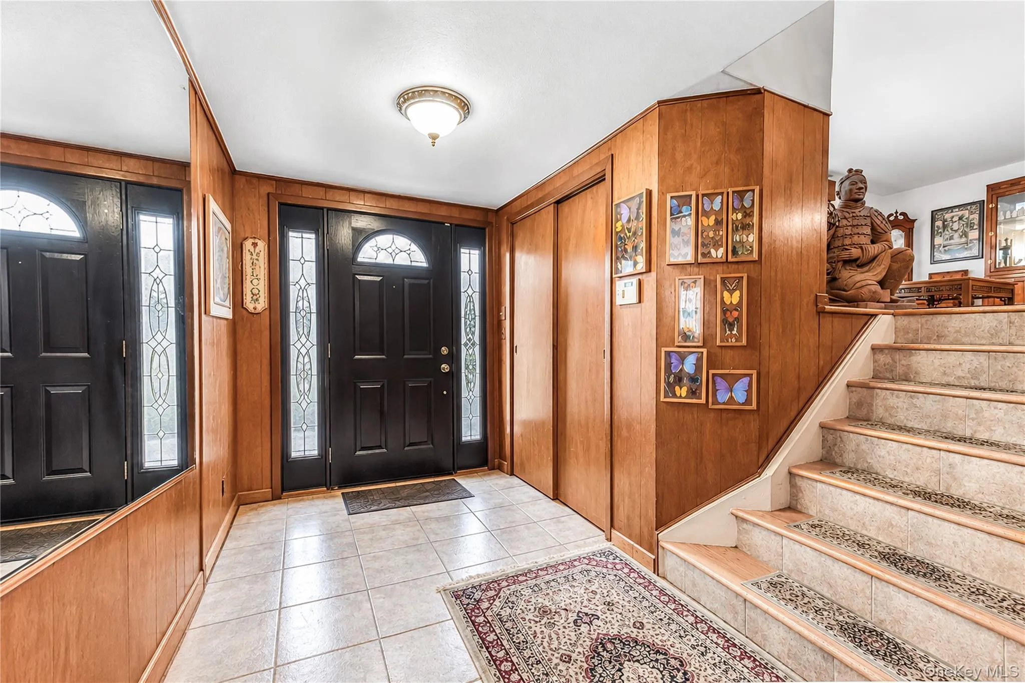 Foyer featuring a black entry door with decorative glass sidelights, tile flooring, wood paneling, and a staircase with an integrated runner Foyer featuring a black entry door with decorative glass sidelights, tile flooring, wood paneling, and a staircase with an integrated runner