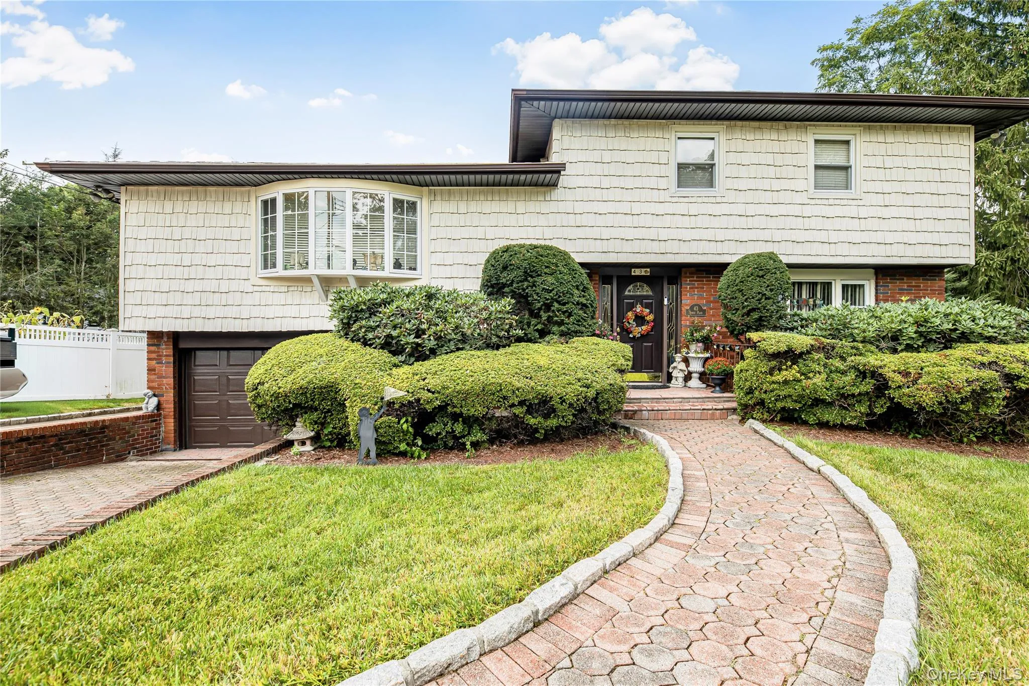 Split-level home featuring a brick paver walkway, mature shrubbery, a bay window, and a brown garage door Split-level home featuring a brick paver walkway, mature shrubbery, a bay window, and a brown garage door