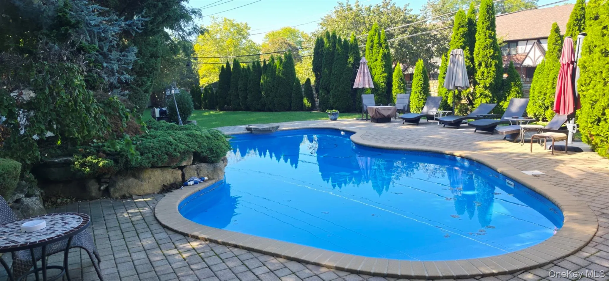 Freeform swimming pool with a paved patio surround, mature privacy landscaping, and a natural stone retaining wall Freeform swimming pool with a paved patio surround, mature privacy landscaping, and a natural stone retaining wall