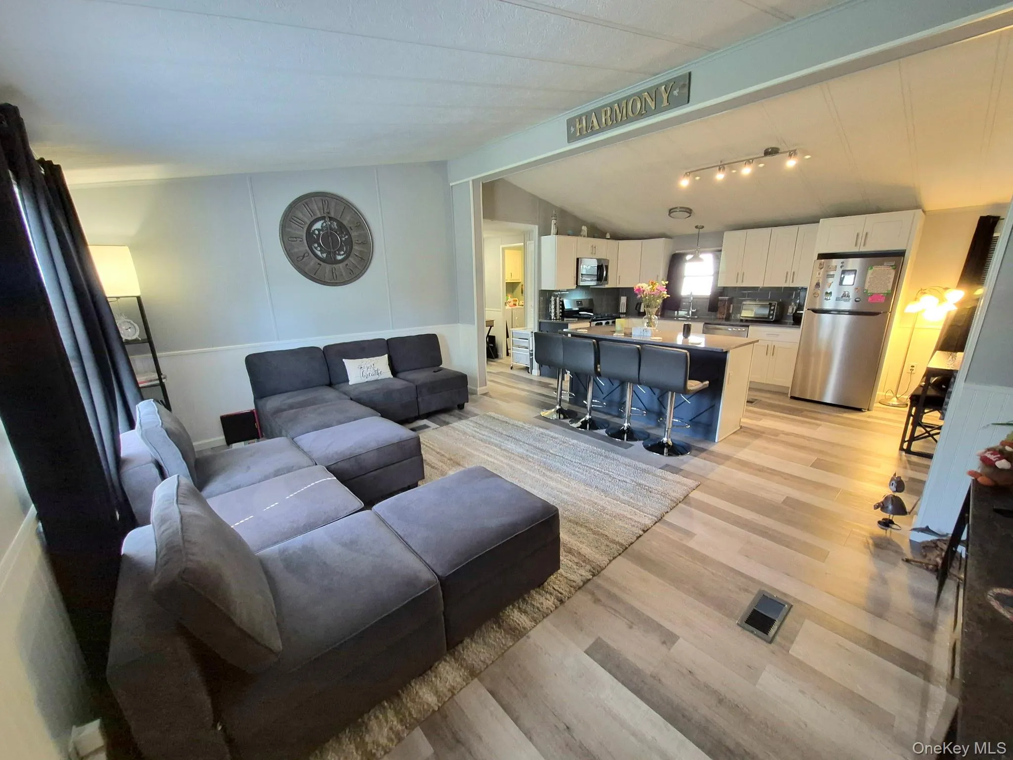 Open-concept living area featuring wood-finish flooring, a kitchen island with seating, stainless steel appliances, white cabinetry, and track lighting Open-concept living area featuring wood-finish flooring, a kitchen island with seating, stainless steel appliances, white cabinetry, and track lighting
