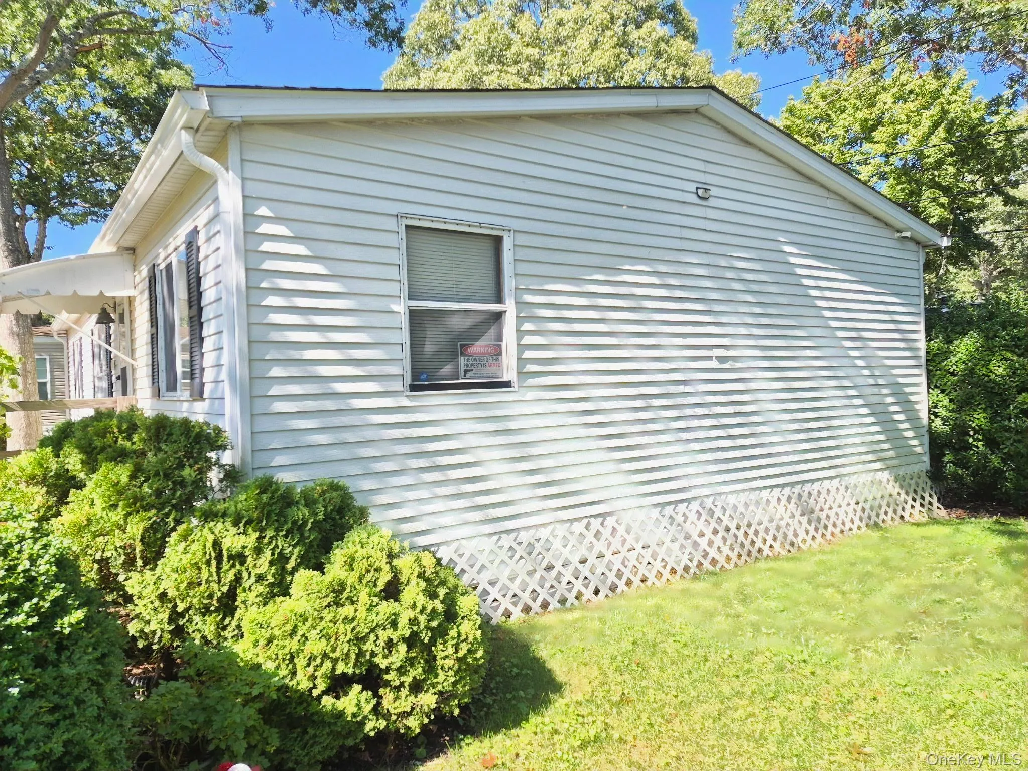 White vinyl siding exterior with an awning over the entrance, featuring green shrubbery and a partial lawn White vinyl siding exterior with an awning over the entrance, featuring green shrubbery and a partial lawn
