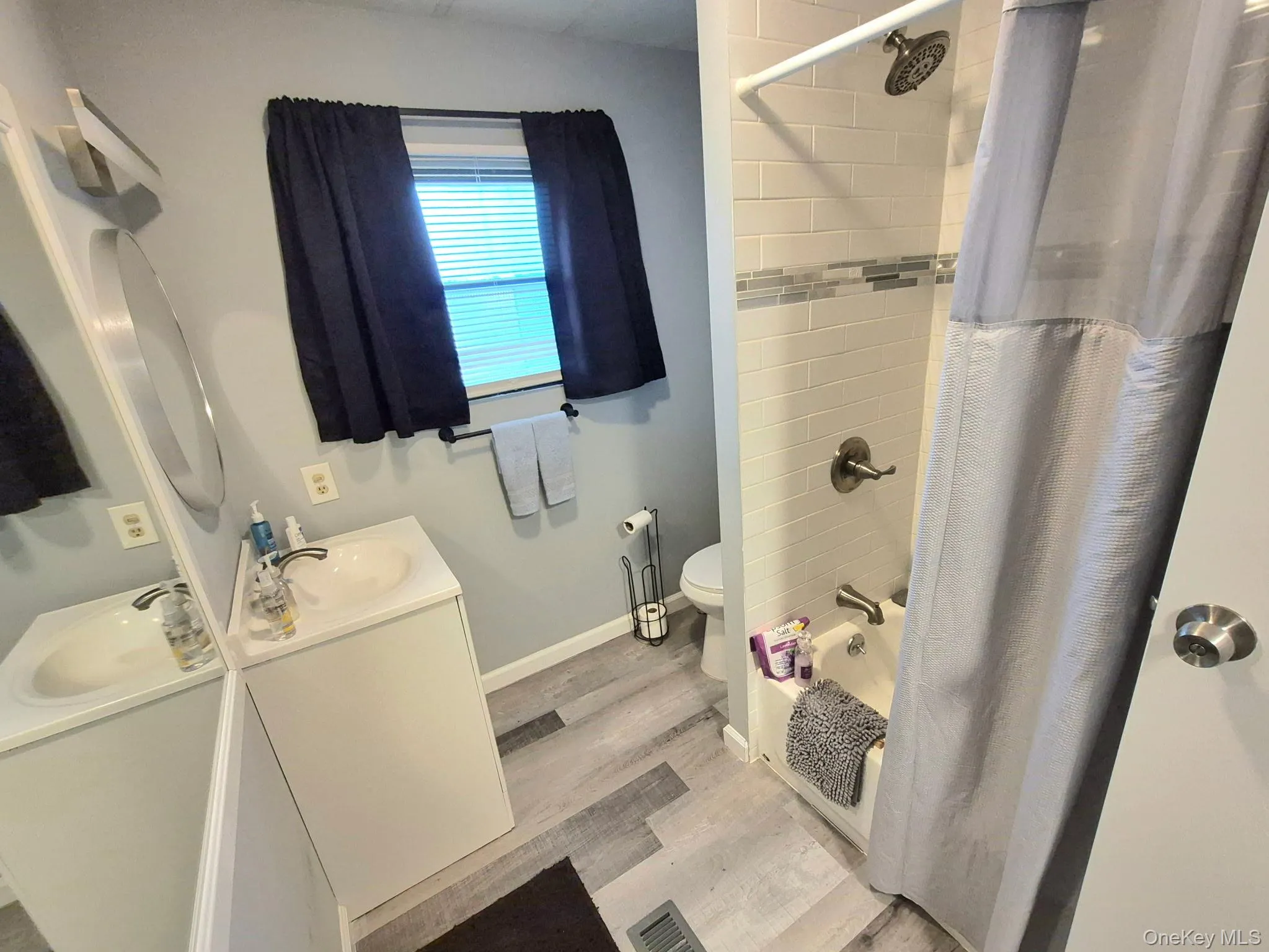 Bathroom featuring wood-finish flooring, a white vanity with an integrated sink, a built-in tub with a shower, and subway tile surround with a mosaic accent Bathroom featuring wood-finish flooring, a white vanity with an integrated sink, a built-in tub with a shower, and subway tile surround with a mosaic accent