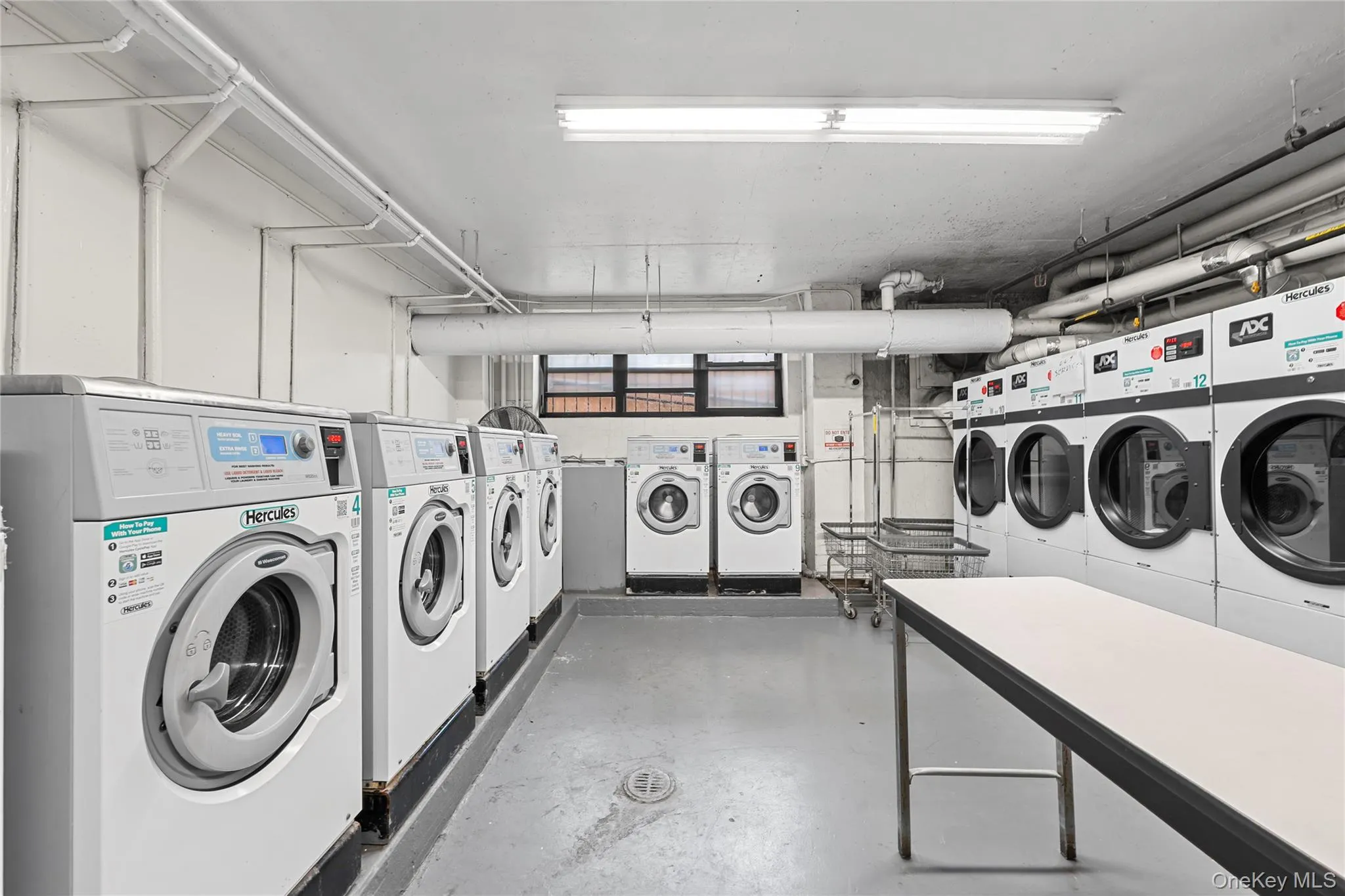 On-site laundry room featuring multiple front-loading washers and dryers, a dedicated folding table, and durable concrete flooring On-site laundry room featuring multiple front-loading washers and dryers, a dedicated folding table, and durable concrete flooring