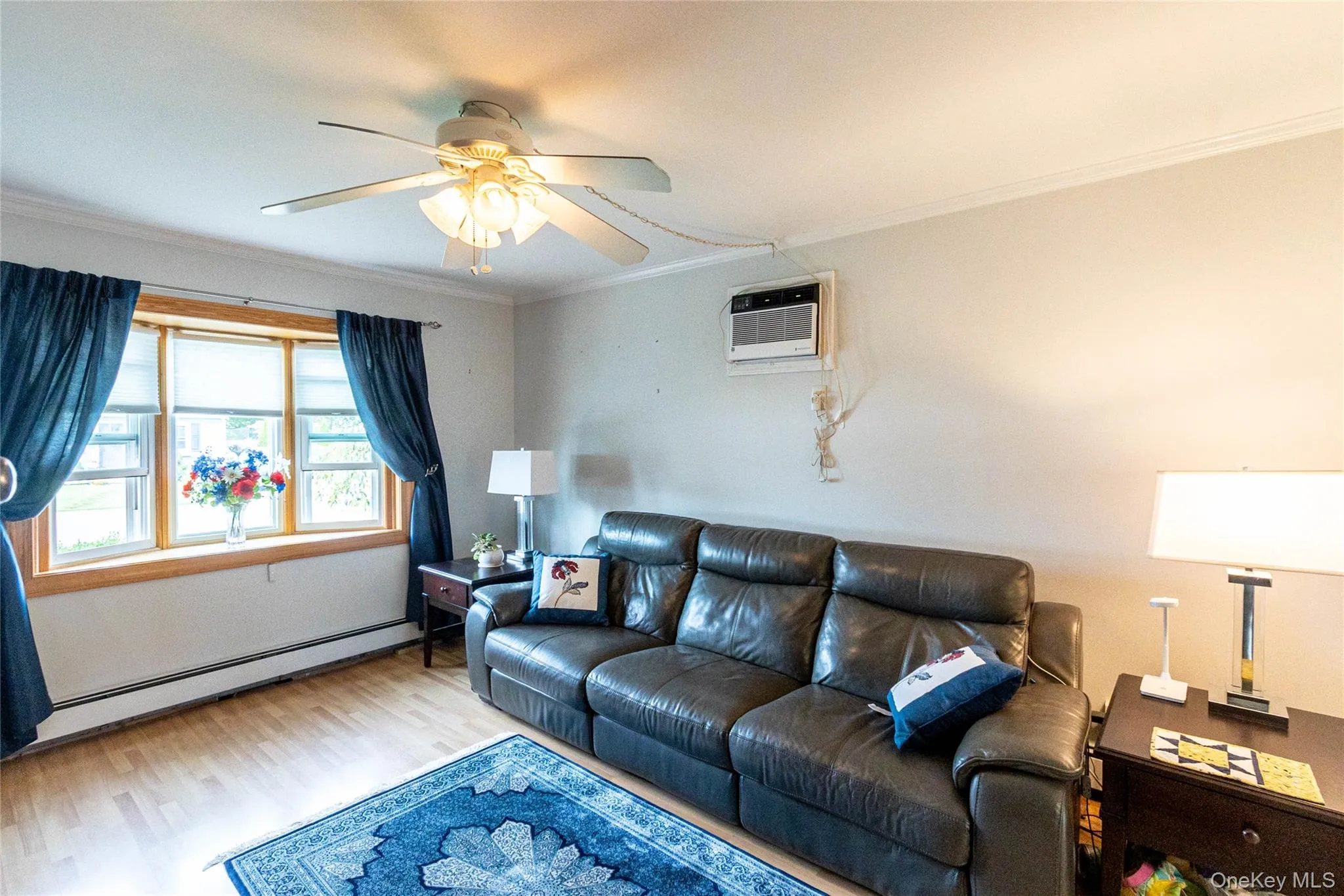 Light wood-finish flooring, ceiling fan with integrated lighting, window-mounted air conditioning unit, and bay window with wood trim Light wood-finish flooring, ceiling fan with integrated lighting, window-mounted air conditioning unit, and bay window with wood trim